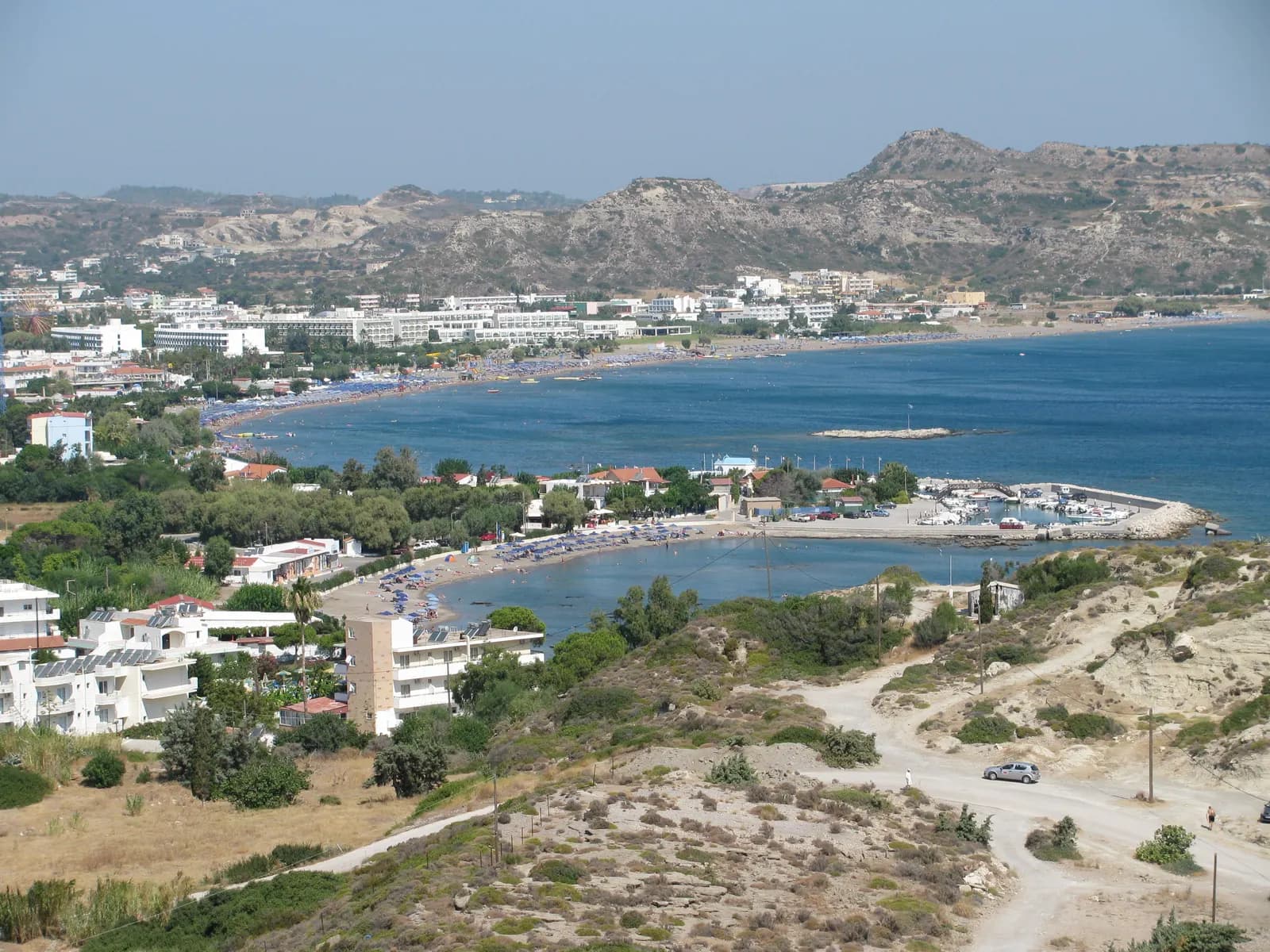 Faliraki bay seen from a hillside, with hotels and apartments along the sandy beach, a small harbour on the right and dry hills behind