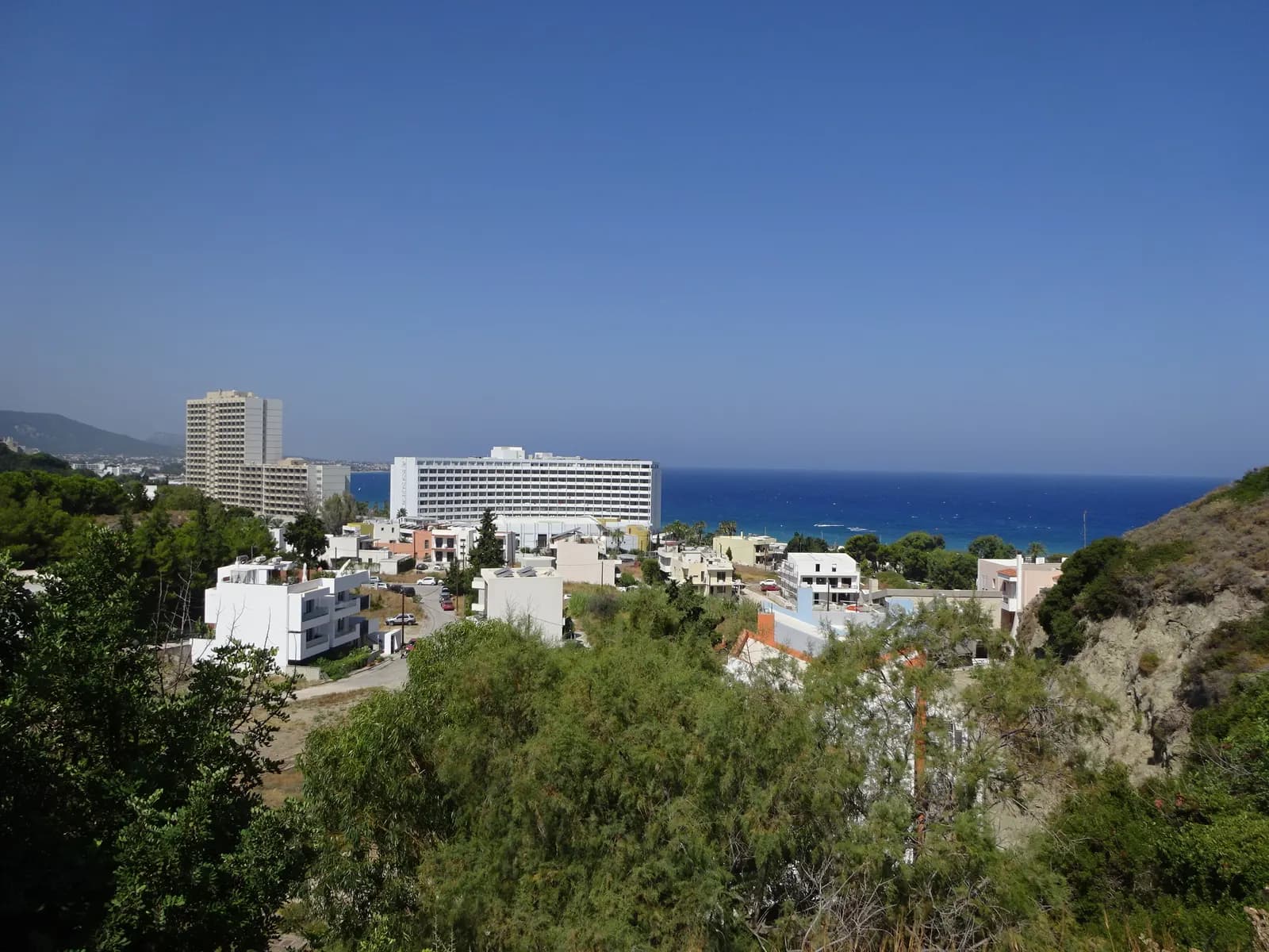 Large resort hotels on the Ixia coast near Ialysos, seen over trees and low white houses, with the blue sea behind