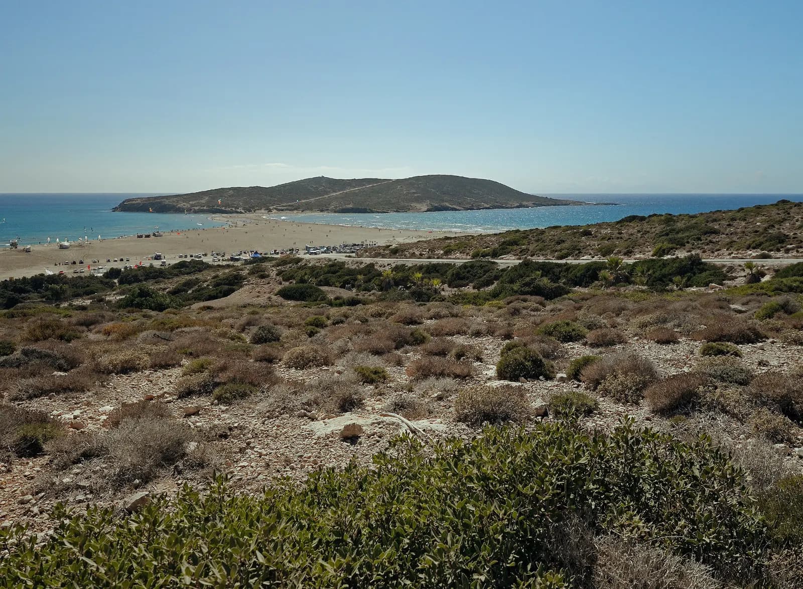 Prasonisi at the southern tip of Rhodes, a sand isthmus with a kite beach joining the island to a hilly islet, seen across low scrub