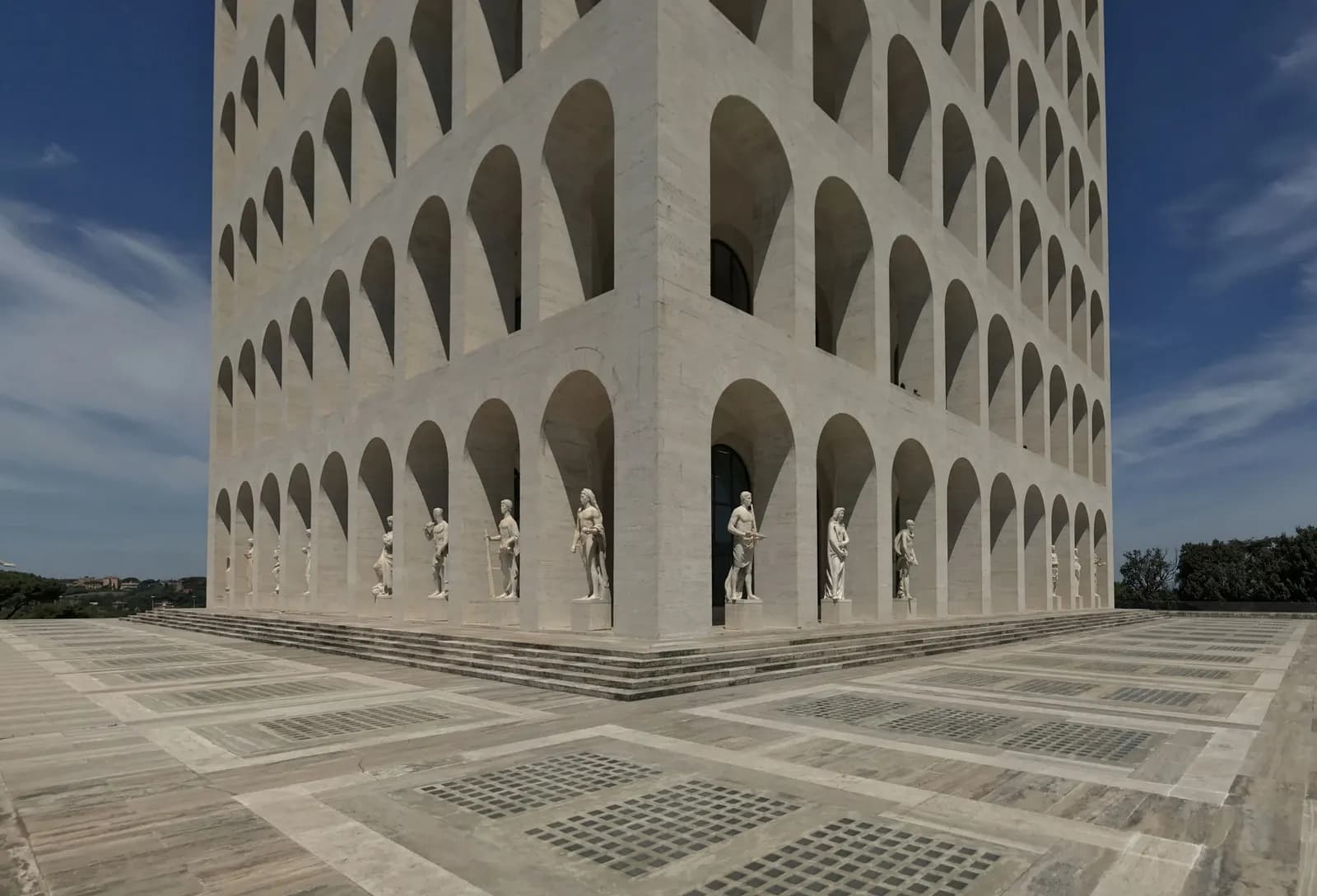 The white arched Palazzo della Civiltà Italiana in EUR, with statues in the ground-floor arches and a wide paved terrace.