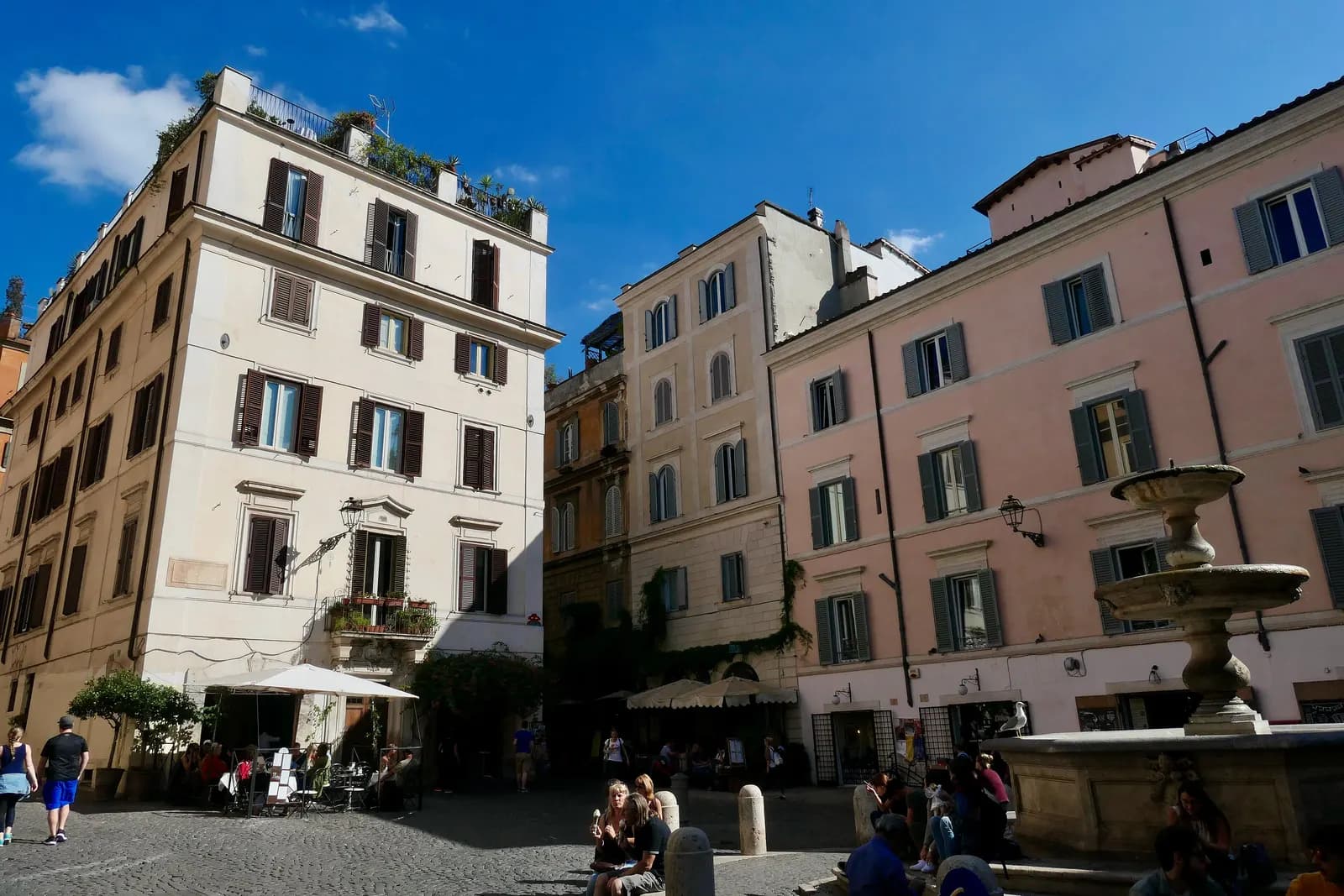 The fountain in Piazza della Madonna dei Monti, with people on its steps, café tables and cream and pink houses under a blue sky.