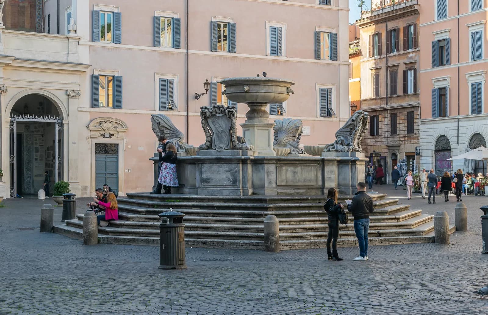The stepped fountain in Piazza di Santa Maria in Trastevere, with pink and ochre houses and café umbrellas behind it.