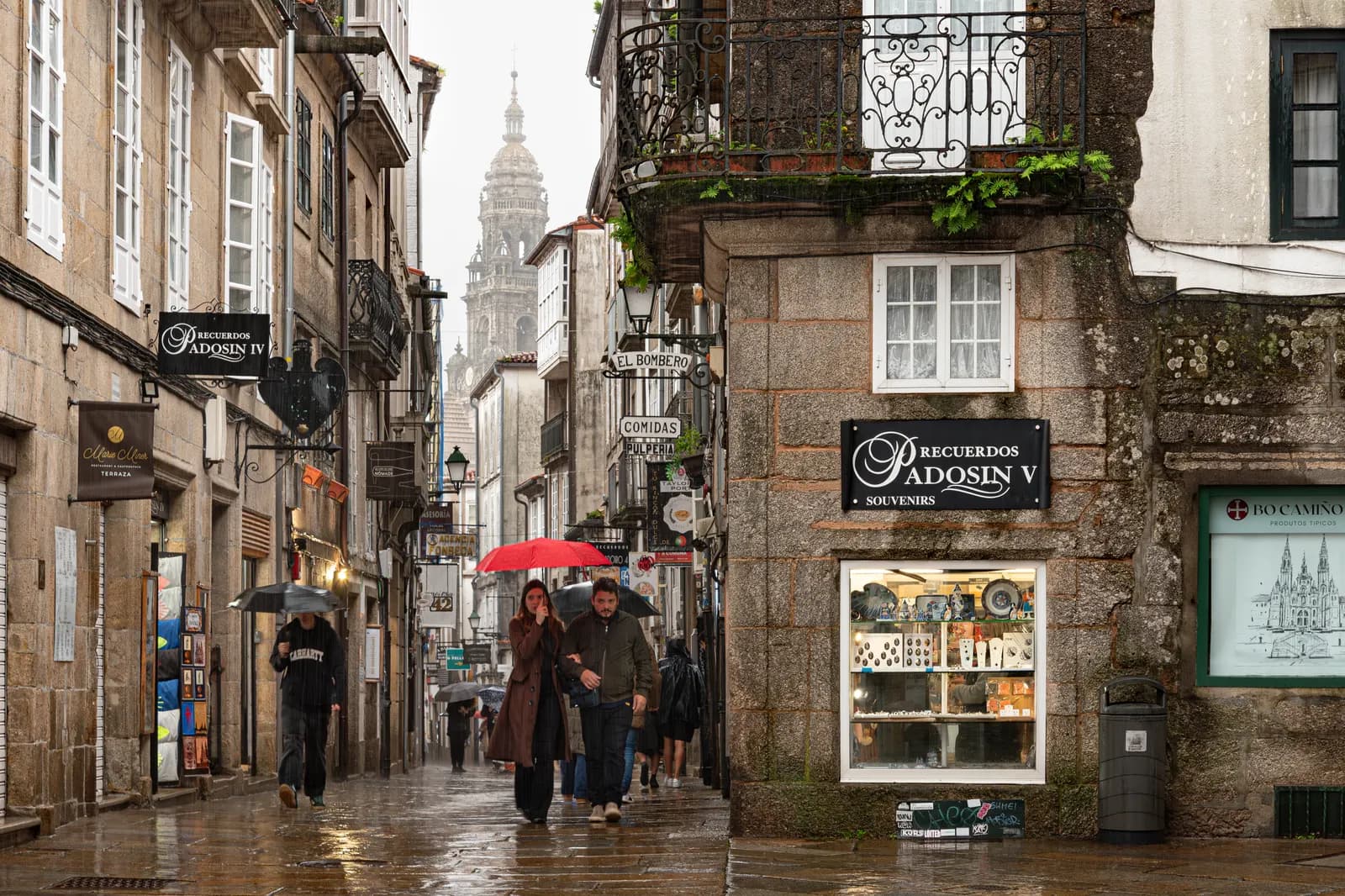 A granite street in Santiago's old town on a rainy afternoon, with souvenir shops, walkers under umbrellas and a cathedral tower at the far end.