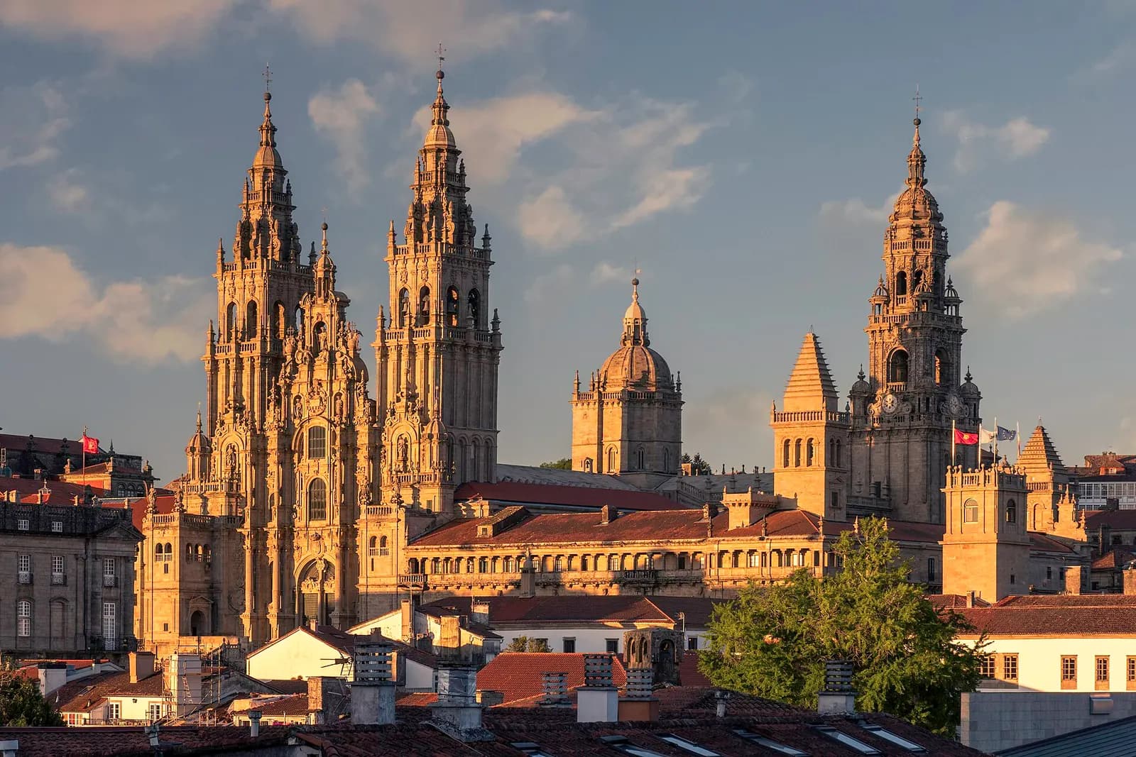 The towers of Santiago de Compostela cathedral lit by late sun above the old town, seen from the Alameda park under a cloudy sky.