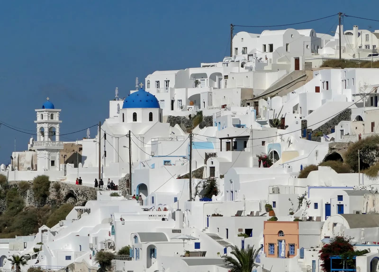 White houses of Imerovigli stepped down the hillside below a blue domed church and a white bell tower, under a clear blue sky