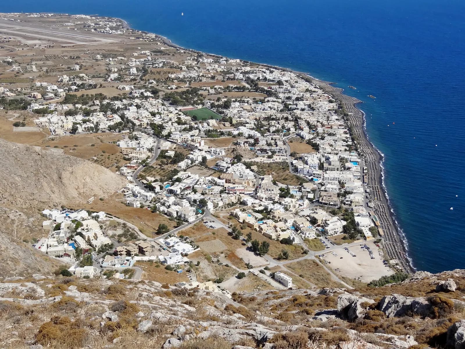 Kamari seen from the mountainside above, with the town, the long dark beach lined with umbrellas and the airport runway in the distance