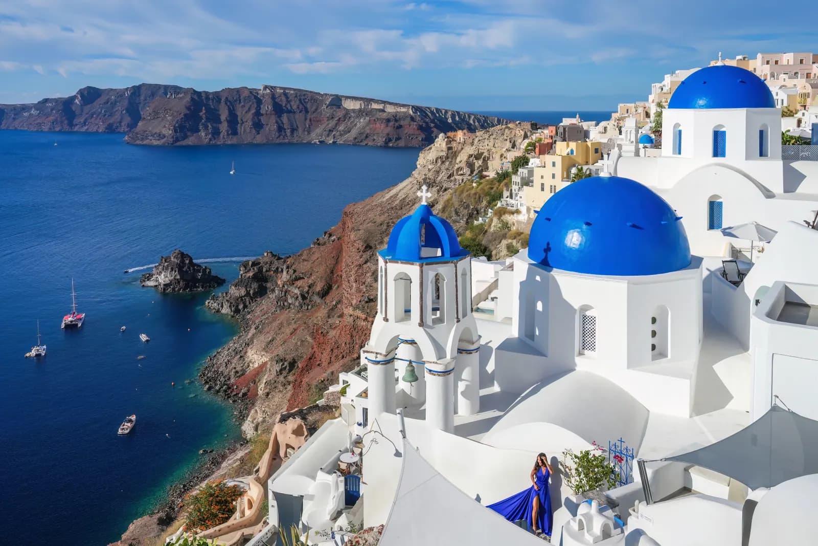 Blue domed churches and a bell tower in Oia on the caldera rim above red cliffs, with boats anchored in the bay below and a woman in a blue dress on a terrace