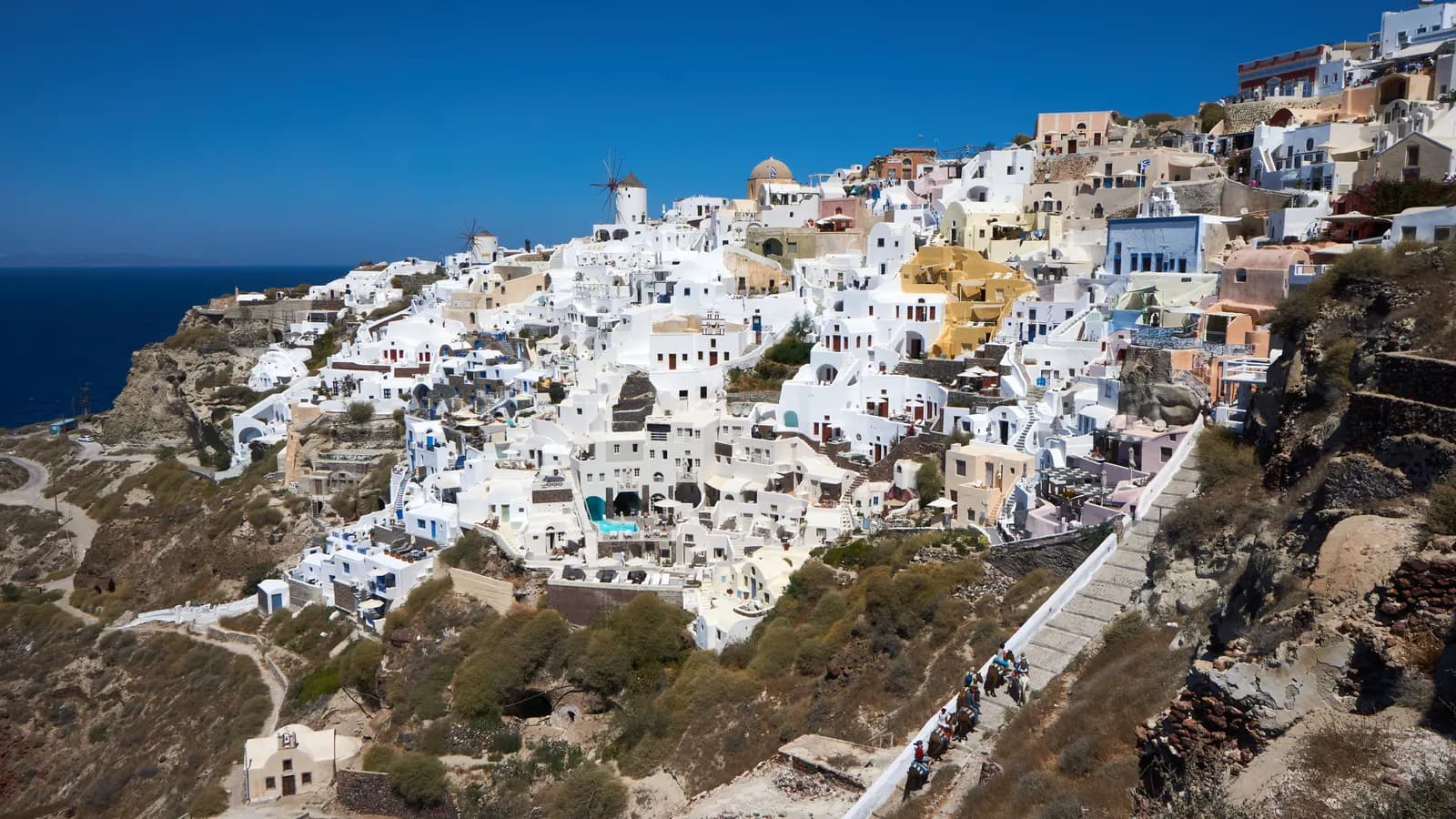 The white and pastel houses of Oia spread across the hillside with windmills on the ridge, a stepped path running down the slope and the sea behind