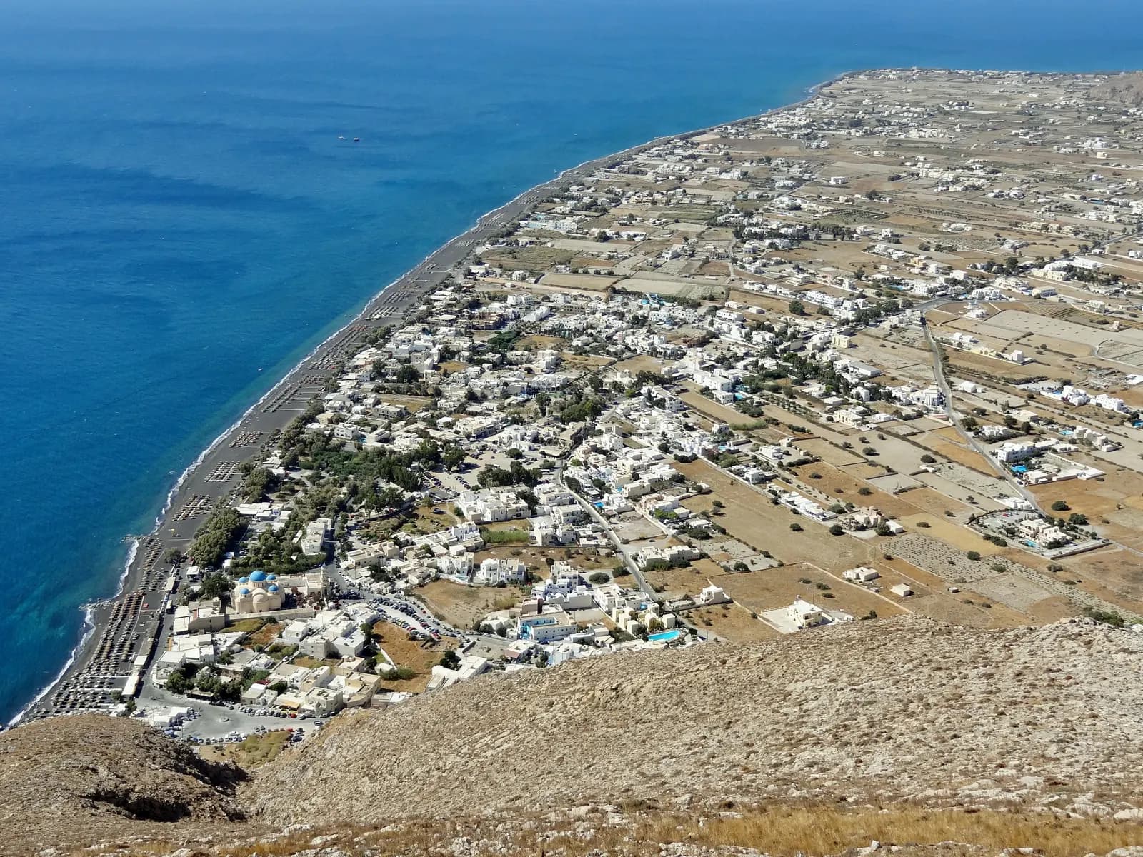 Perissa seen from the mountain above, with its blue domed church, the long dark beach lined with umbrellas and the flat plain along the coast