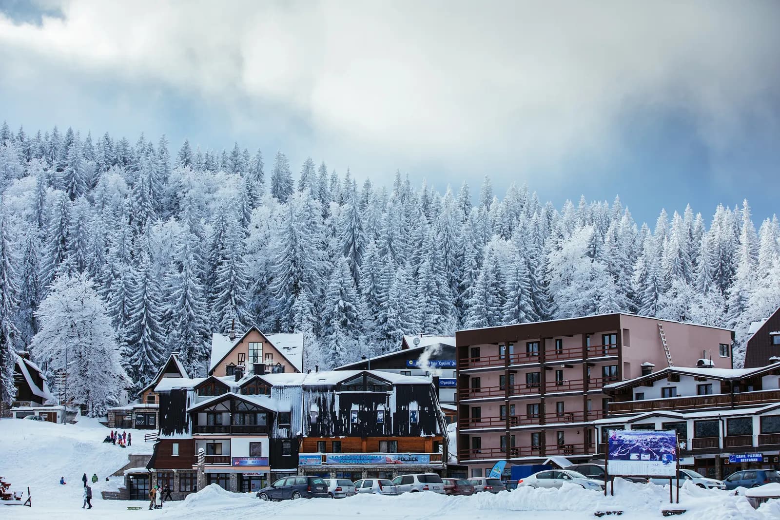 Hotels and chalets below the snow-covered spruce forest at the Jahorina ski resort in December, with people on the snow and cars parked in front