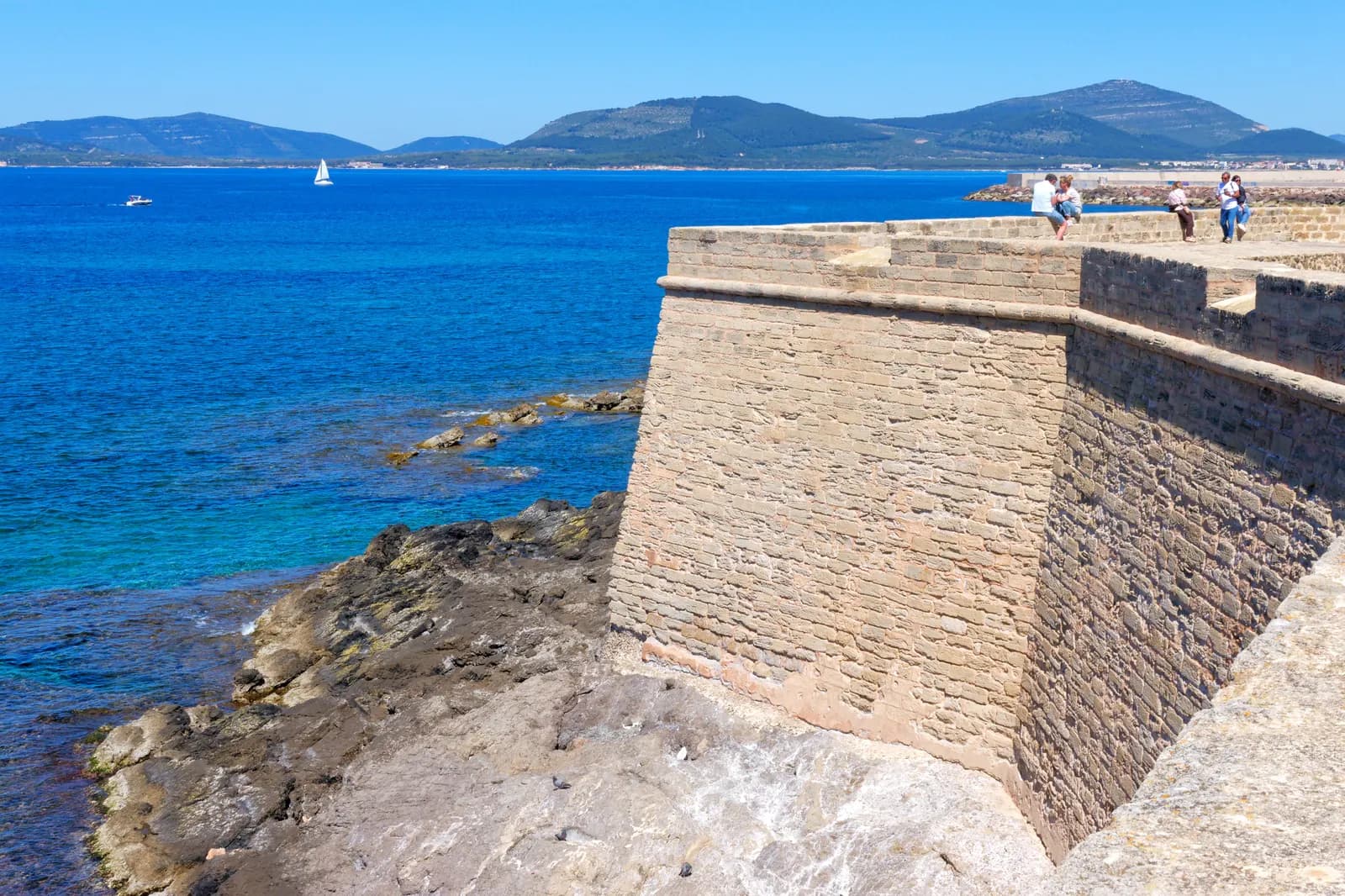 The sandstone sea walls of the old town of Alghero above clear blue water, with people on the ramparts and green hills across the bay