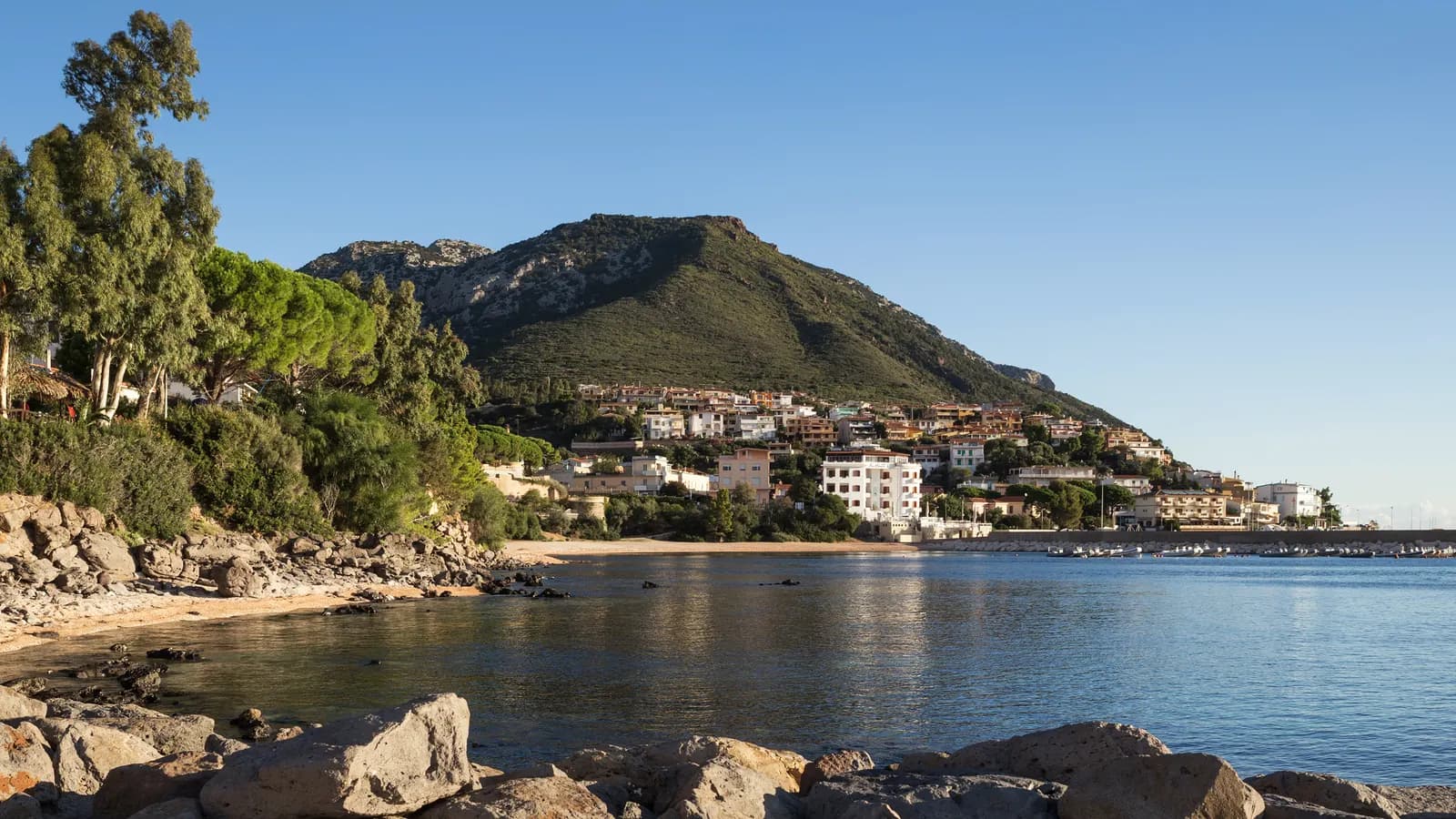 Cala Gonone on the slope of a green limestone mountain above a calm bay, with rocks in the foreground and a small harbour on the right