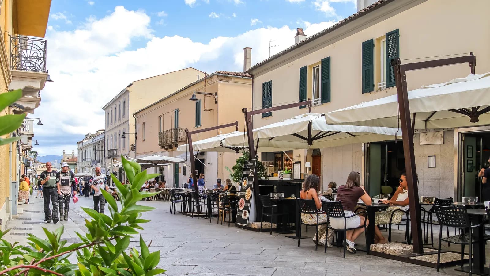 Corso Umberto in the old centre of Olbia, a paved street with cafe tables under white umbrellas and pastel houses with green shutters