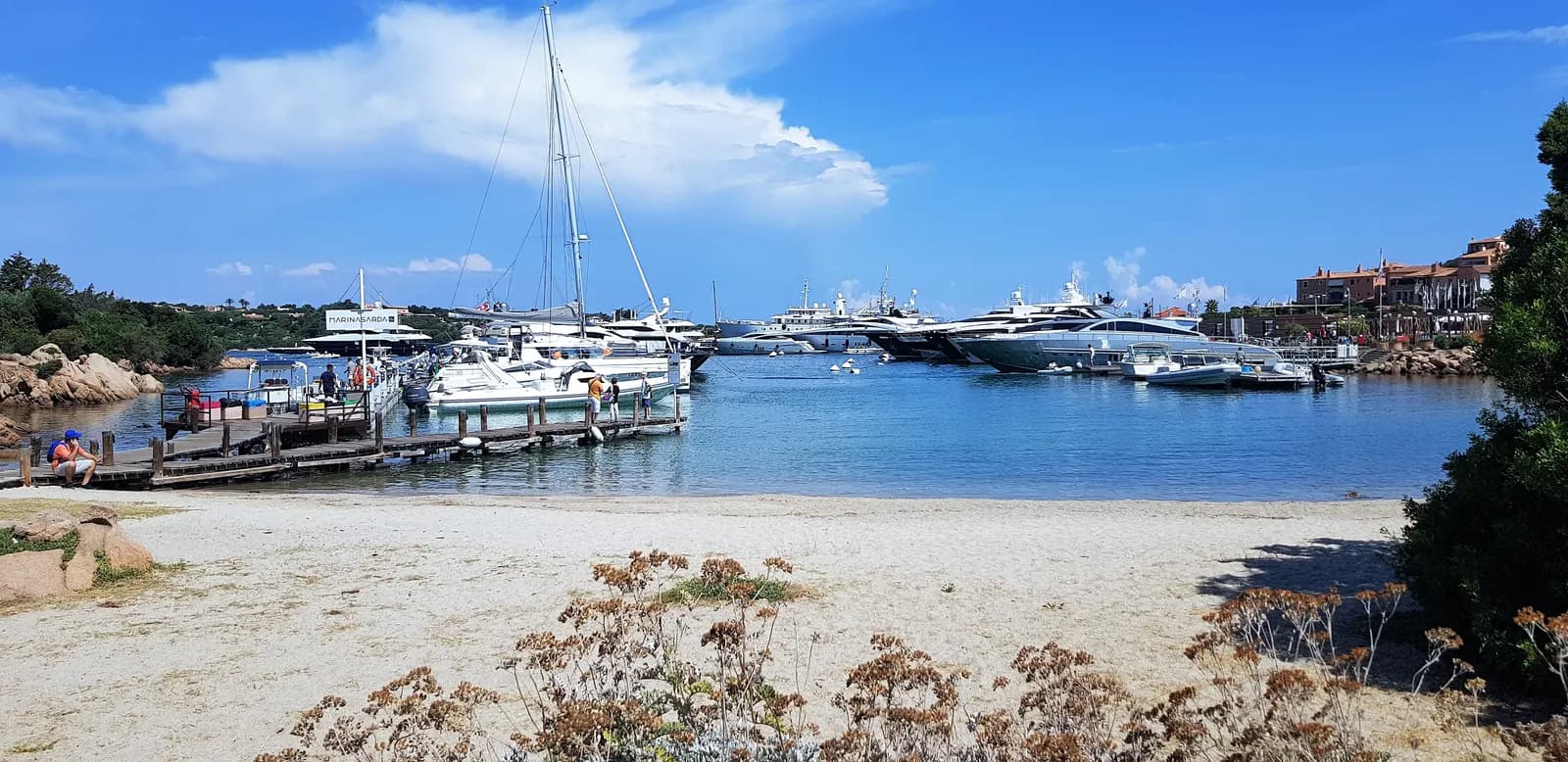 The marina at Porto Cervo seen from the town beach, with motor yachts and a sailing boat moored in a calm bay