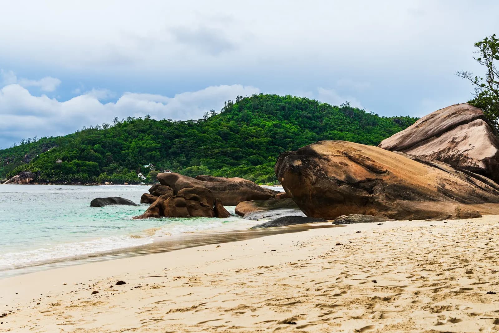 The public beach at Baie Lazare in the south-west of Mahé, with large granite boulders at the water's edge and a wooded hill behind under a cloudy sky