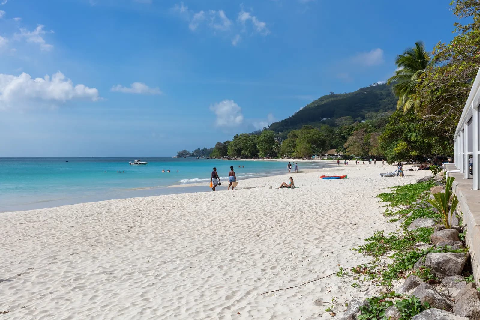 The white sand beach at Beau Vallon on the north-west coast of Mahé, with people walking by the water, a boat in the bay and green hills behind