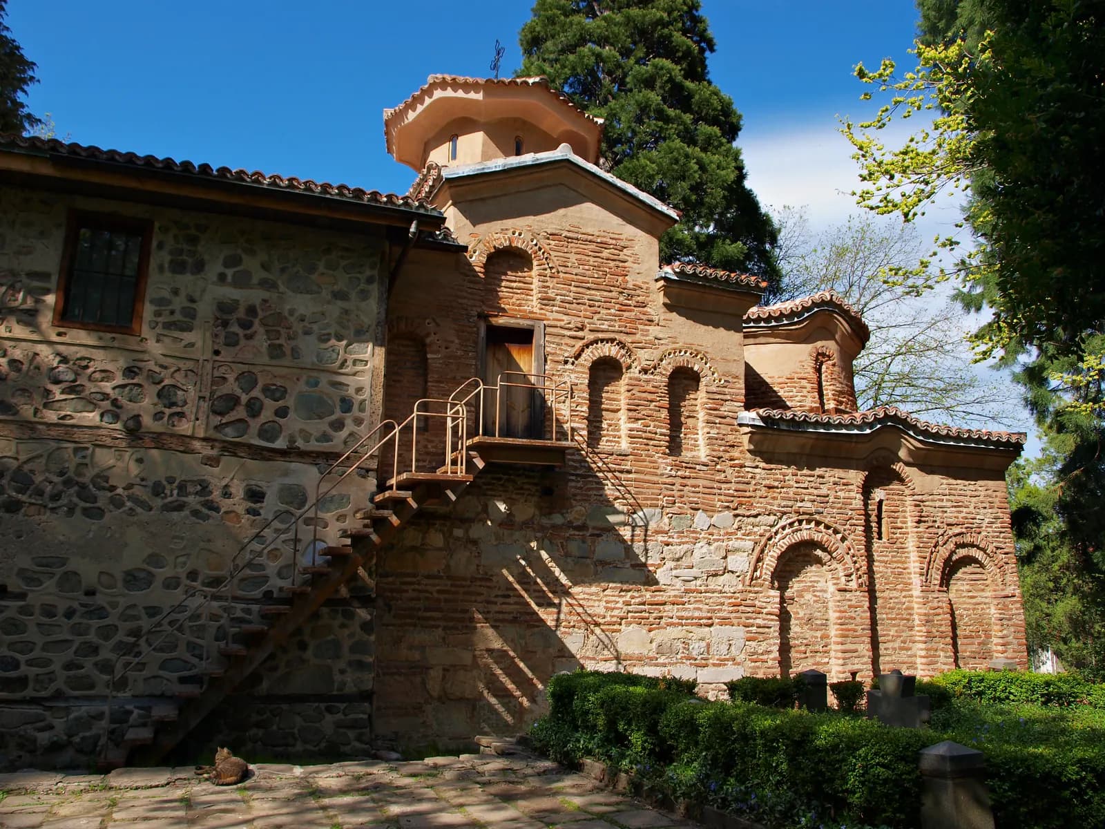 The medieval Boyana Church on the edge of Sofia, with red brick and stone walls, a small drum dome and an outside staircase, among tall trees in sunshine
