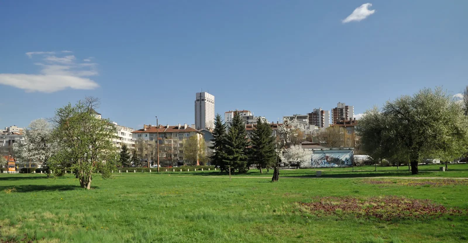 Apartment buildings of Lozenets and a tall white hotel tower seen across the lawns and blossoming trees of South Park in Sofia
