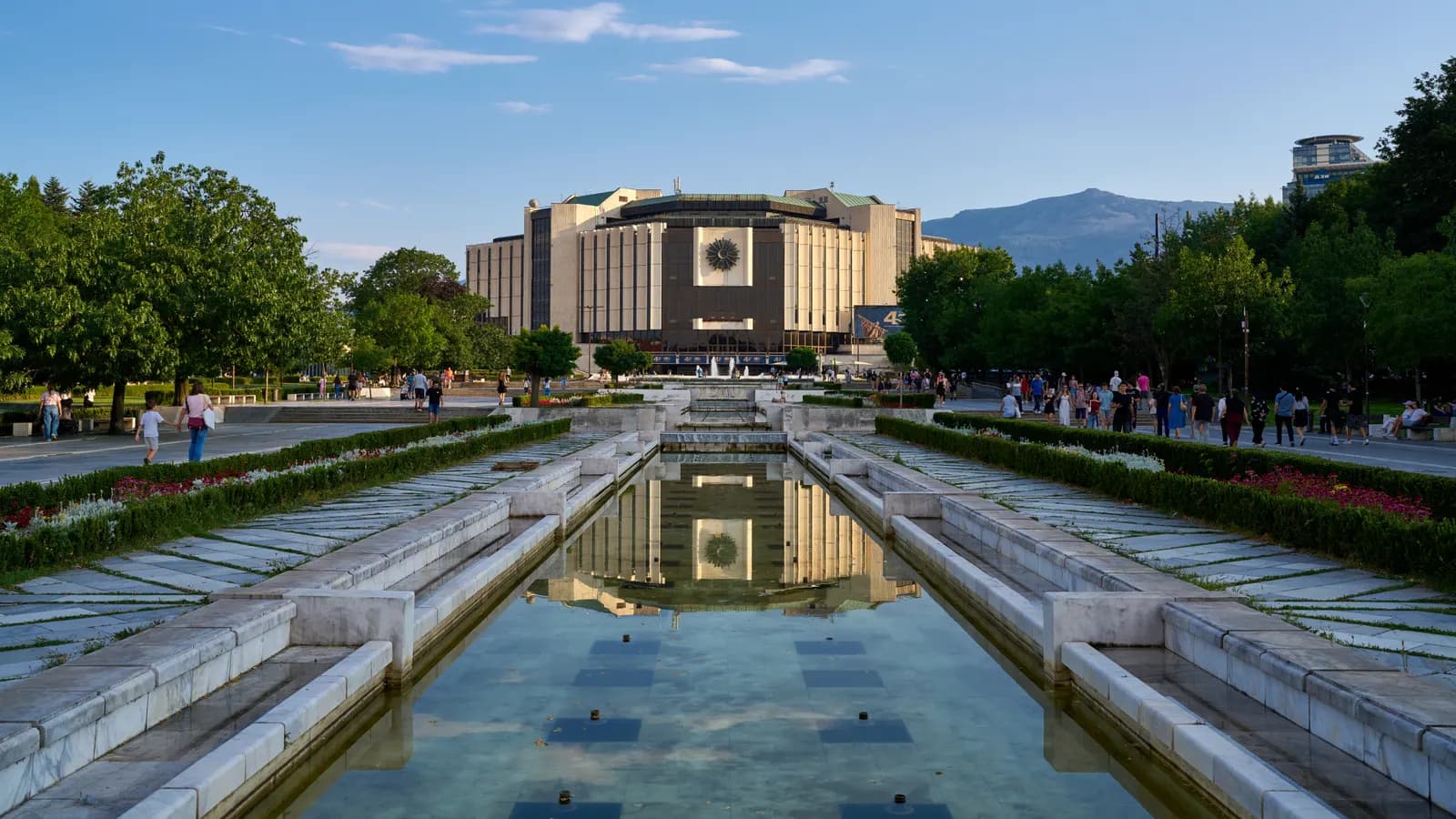 The National Palace of Culture at the southern end of central Sofia, seen along a long fountain pool lined with flower beds and strolling people, with Vitosha mountain behind