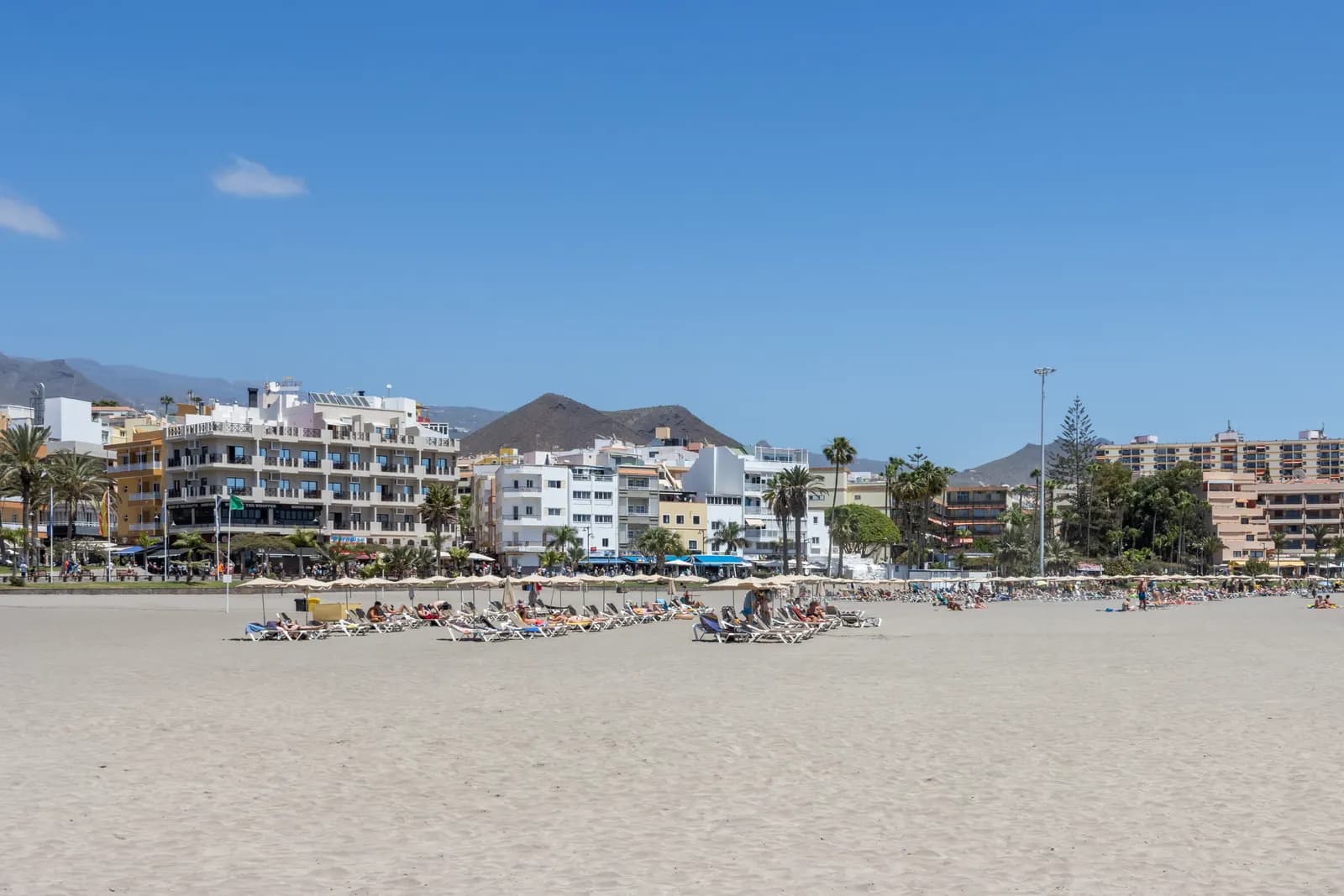 Wide sandy beach at Los Cristianos with sun loungers and a row of apartment blocks and palms behind, a volcanic hill on the skyline