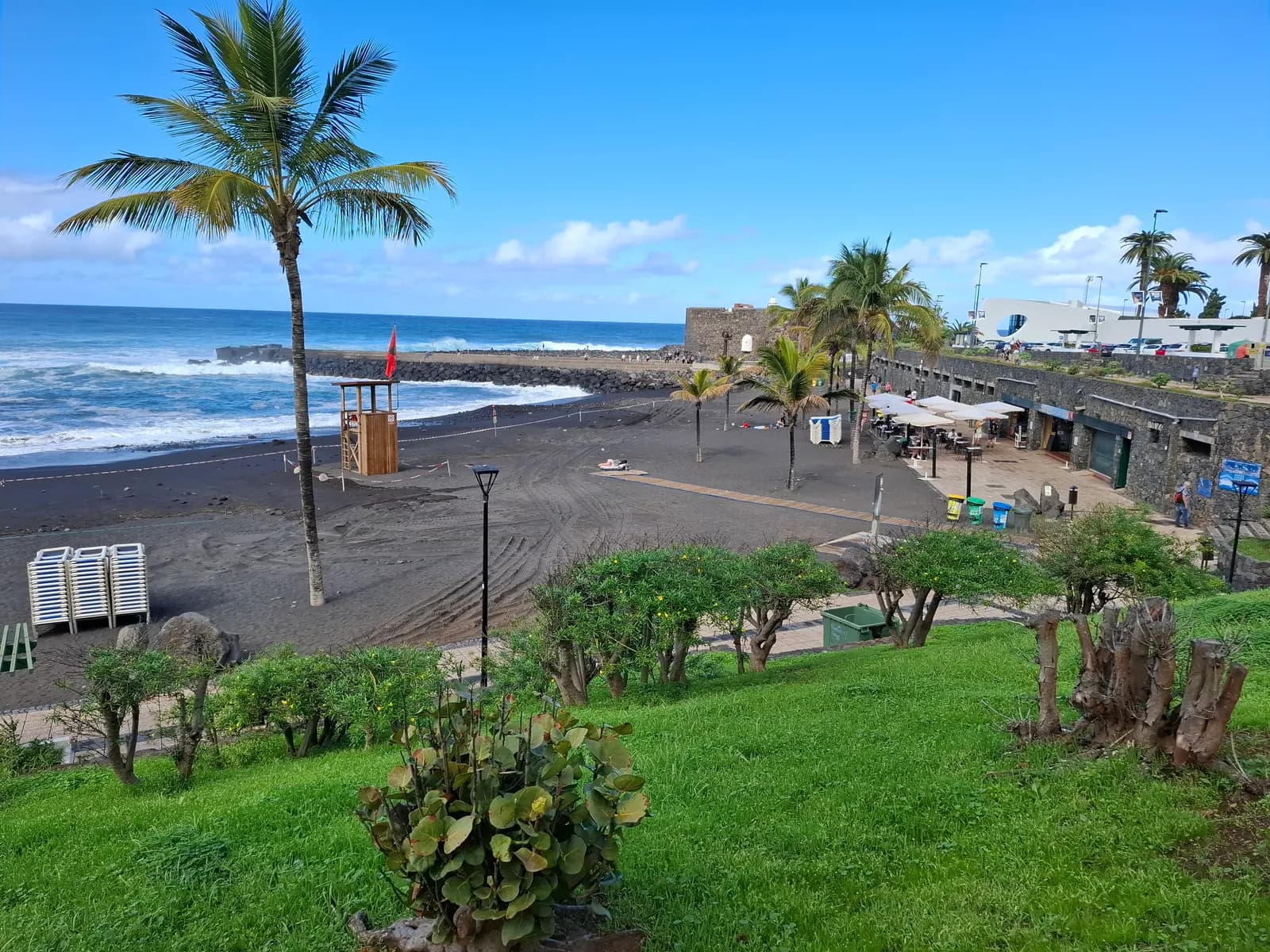 Black sand of Playa Jardín in Puerto de la Cruz with a palm, a lifeguard tower, gardens and the stone Castillo de San Felipe by the sea