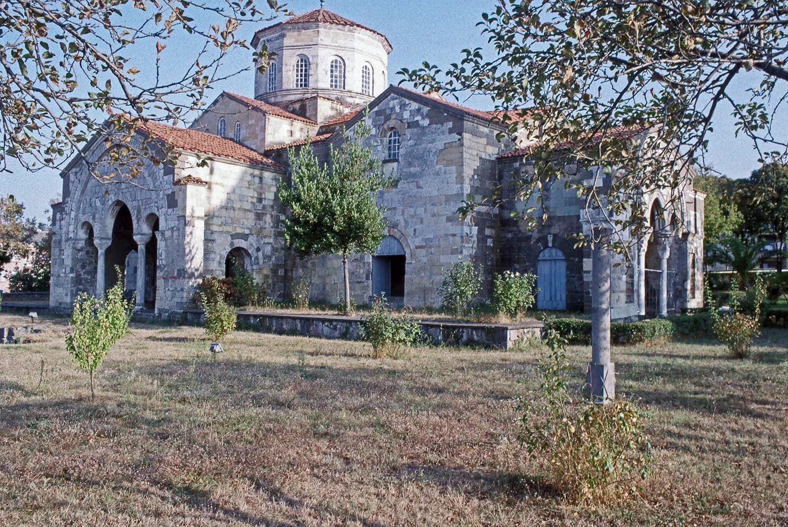 The stone church of Hagia Sophia in Trabzon, with arched porches and a domed drum, behind a lawn with young trees