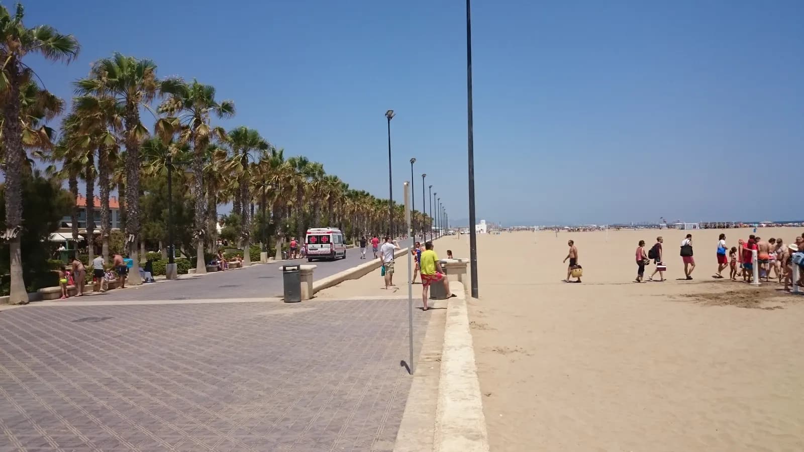 The palm-lined promenade and wide sand of Valencia's city beach on a sunny summer day.