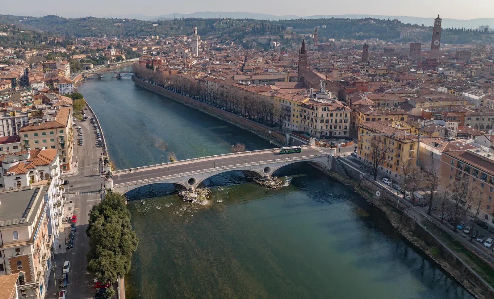 Aerial view of the Ponte della Vittoria over the Adige, with the Borgo Trento riverside road on the left and the towers of the old town and a cypress-covered hill beyond.