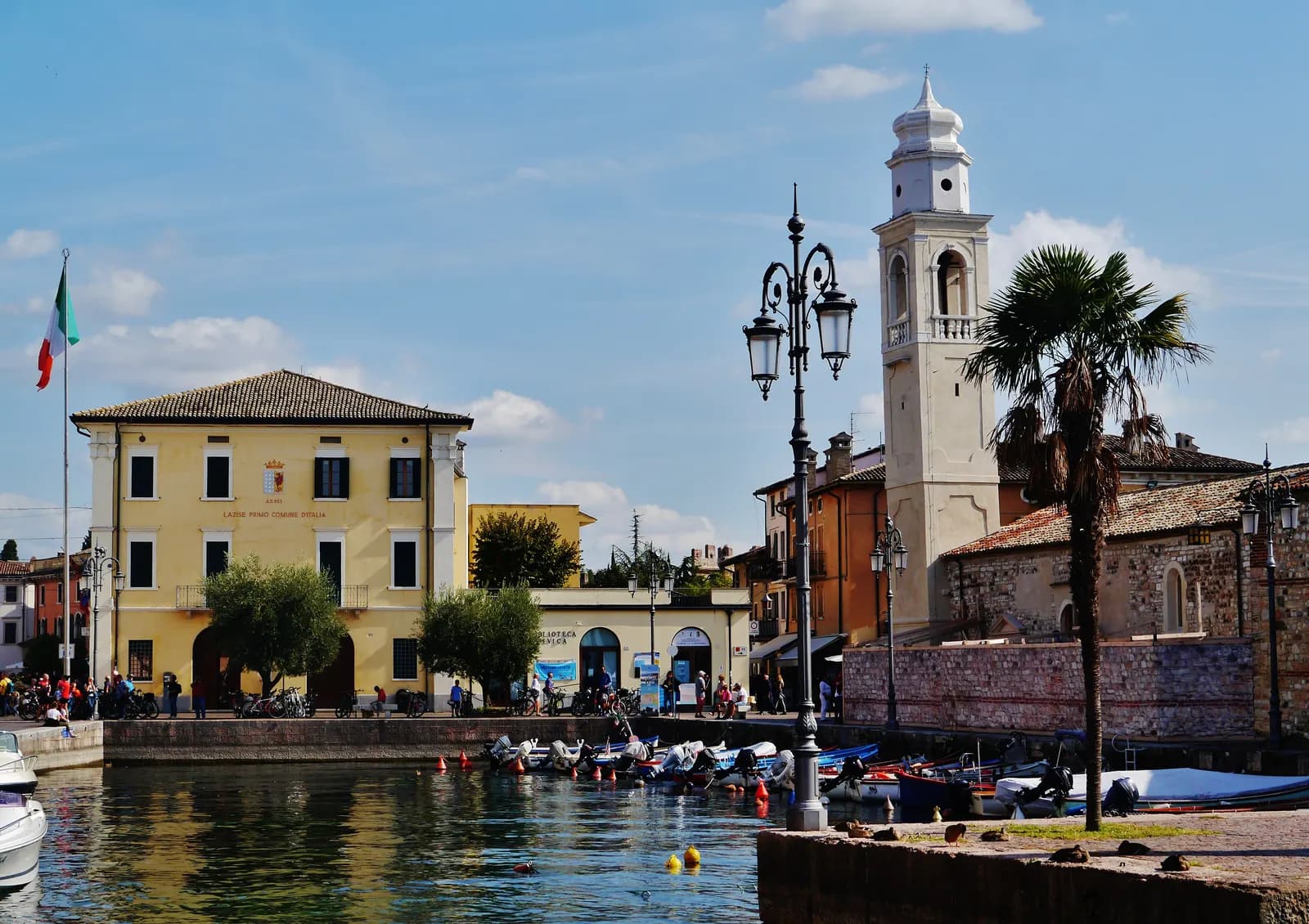 The small harbour of Lazise on Lake Garda, with moored boats, the yellow town hall, the white bell tower of San Nicolò and a palm tree.