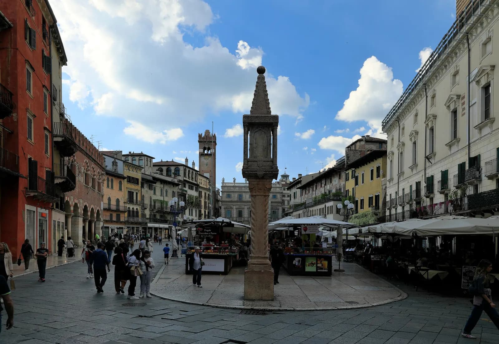 Piazza delle Erbe in Verona, with a carved stone column in front, market stalls and café awnings, and the Torre del Gardello and Palazzo Maffei at the far end.