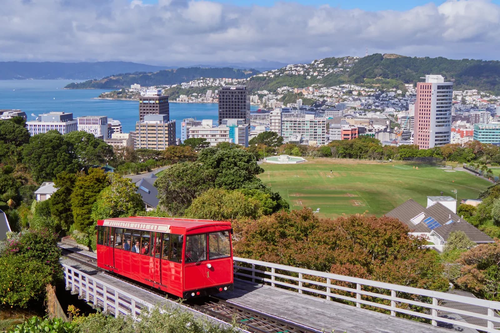 A red Wellington Cable Car on its track above a sports ground, with the city's office towers, the harbour and houses on the hills behind