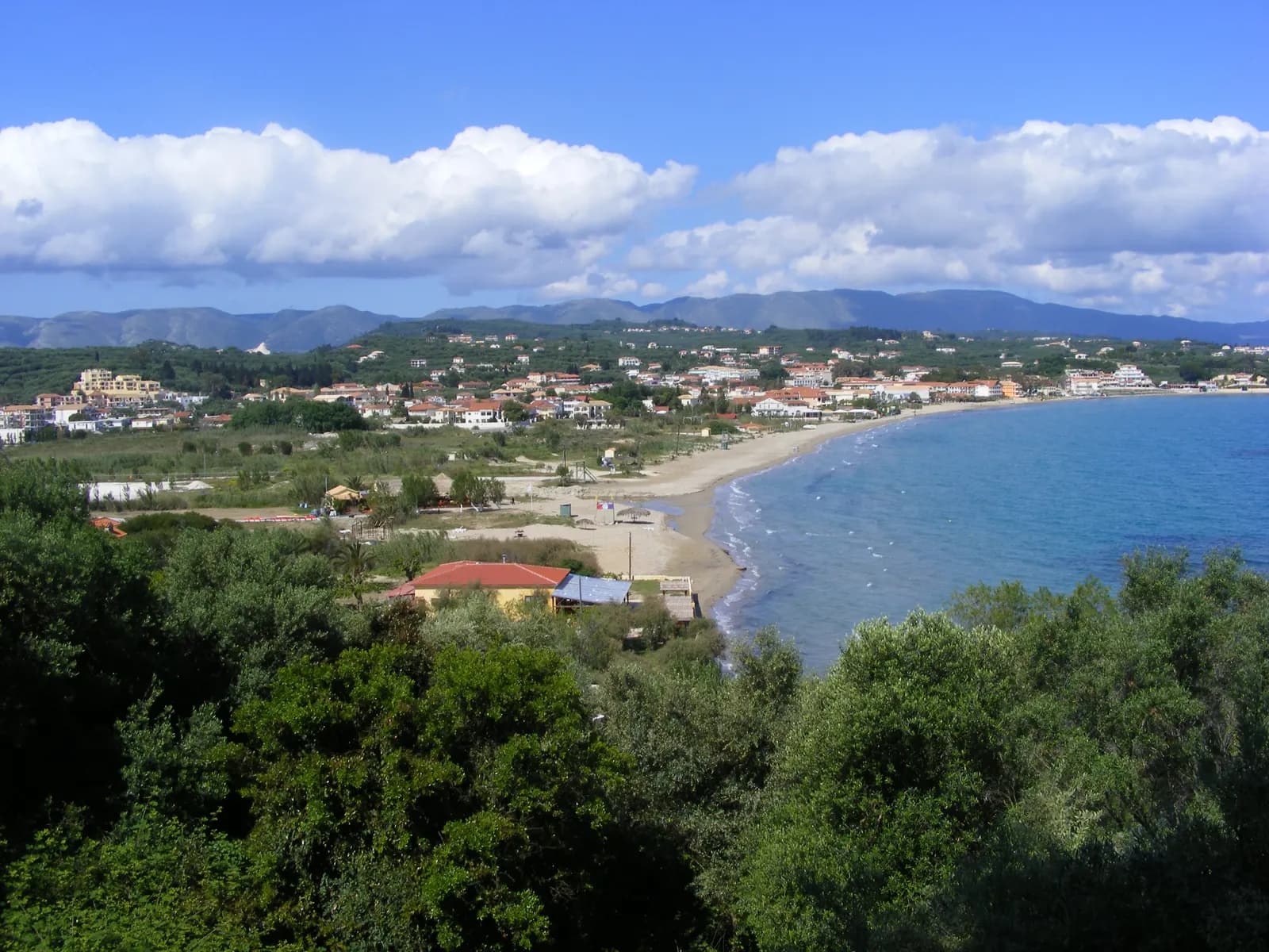 Tsilivi bay on the east coast of Zakynthos seen from a hill, with the curving beach, resort houses among olive trees and mountains under white clouds