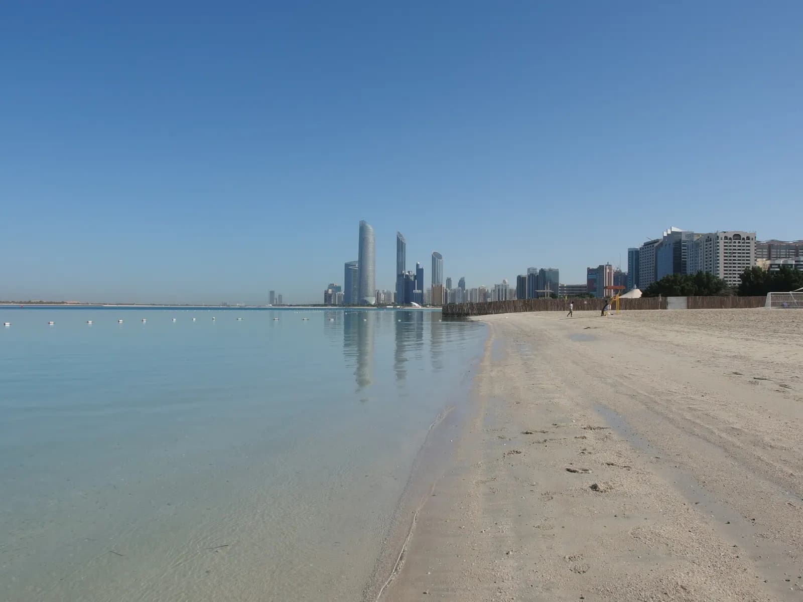 Calm, clear water and a sandy beach on the Abu Dhabi Corniche, with a line of towers along the shore in the distance