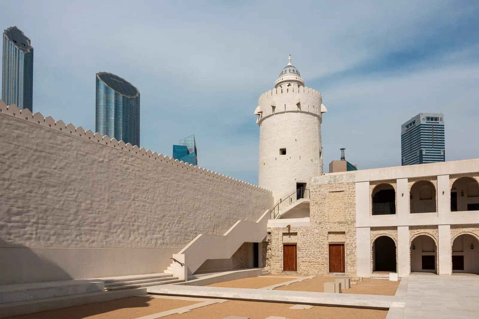 The white round watchtower and crenellated wall of Qasr Al Hosn, the old fort in the centre of Abu Dhabi, with glass towers behind