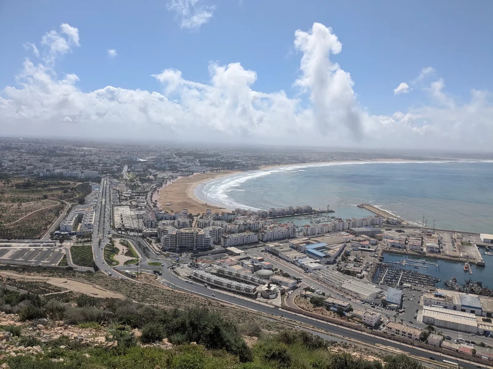 Agadir bay seen from the Kasbah of Agadir Oufella: the curve of the beach, the marina and port below, and the city spreading inland under white clouds