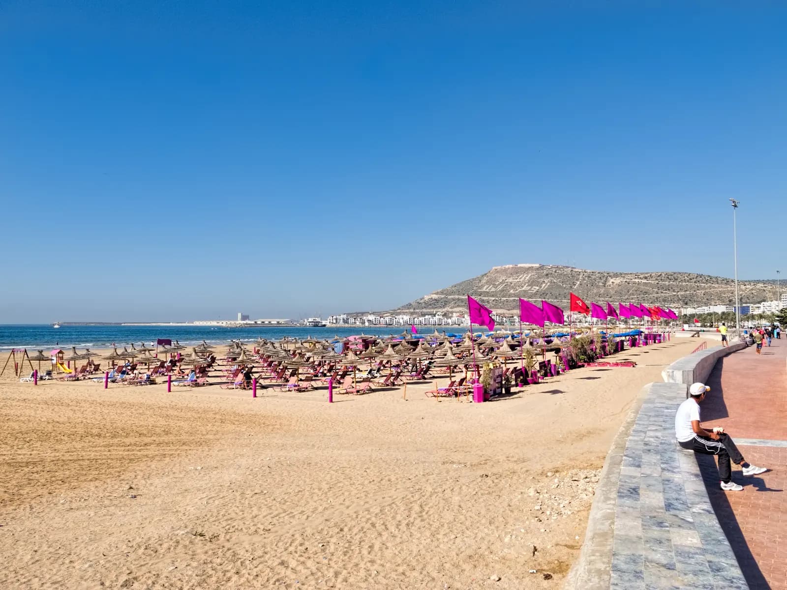 The paved seafront promenade beside Agadir beach, with rows of sunshades and pink flags on the sand and the Kasbah hill in the distance