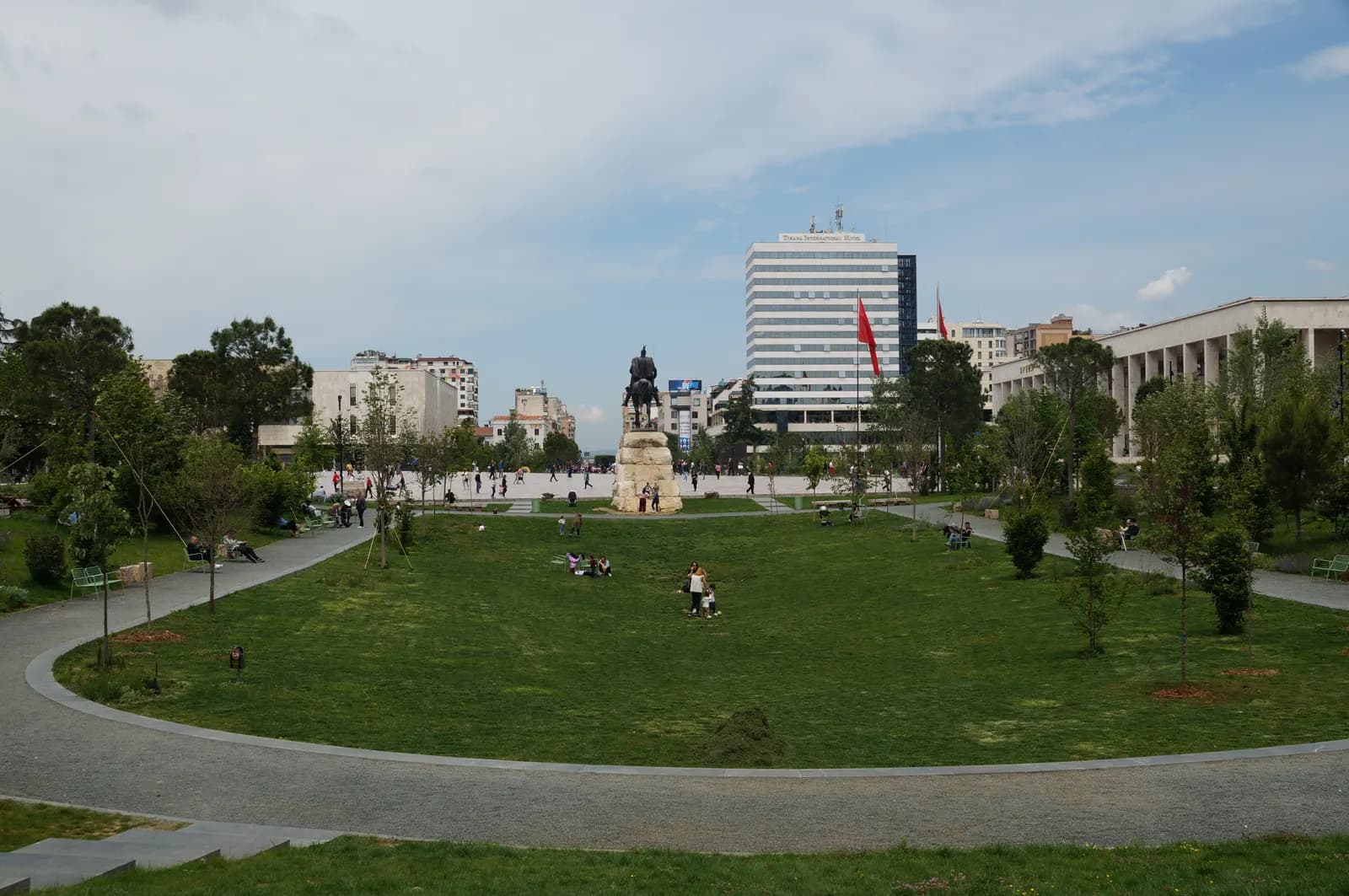 The green lawns and paths of Skanderbeg Square in Tirana, with the Skanderbeg statue, a hotel tower and government buildings behind