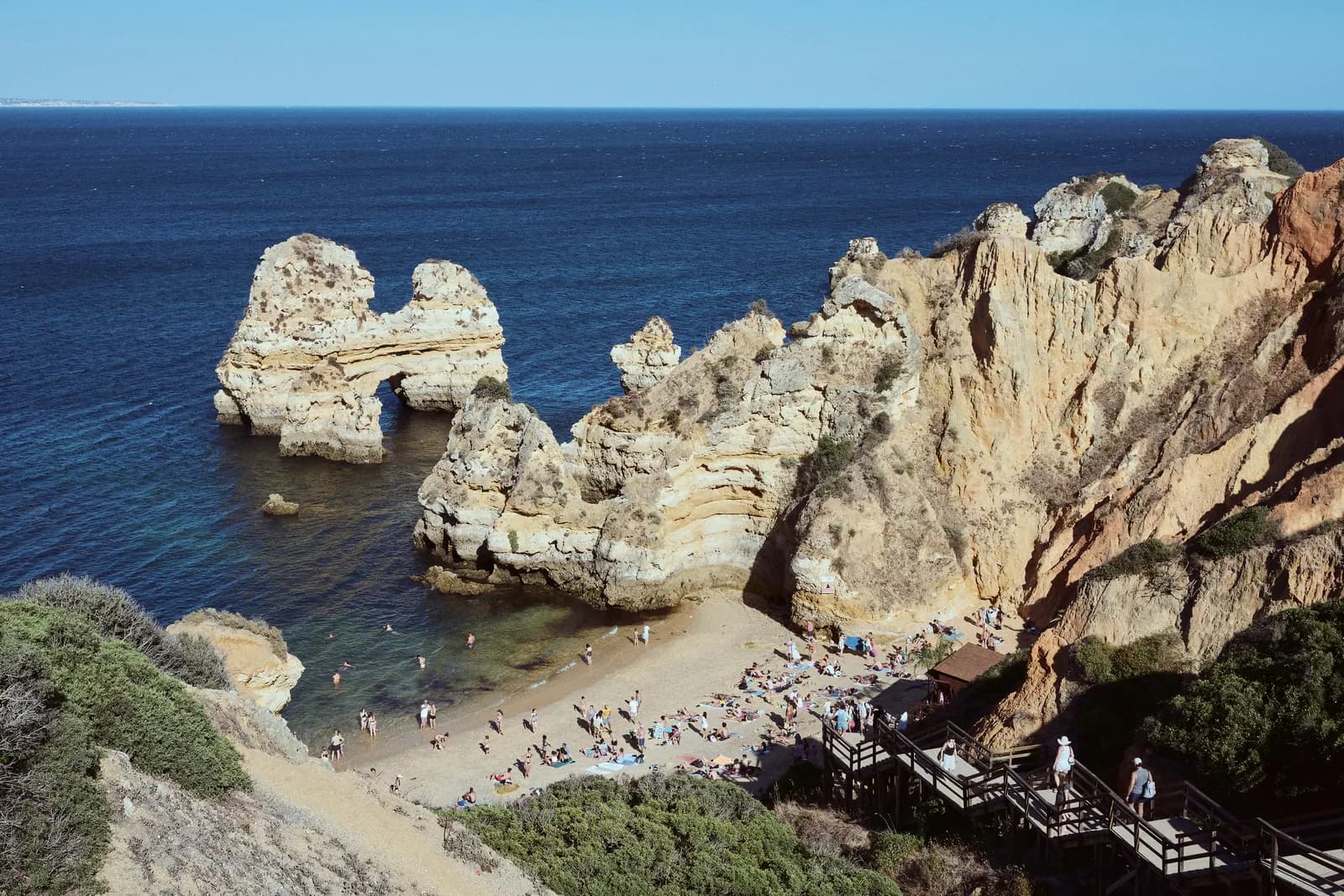 Golden limestone cliffs and sea stacks above a small beach near Lagos, with a wooden stairway down the cliff and the Atlantic beyond