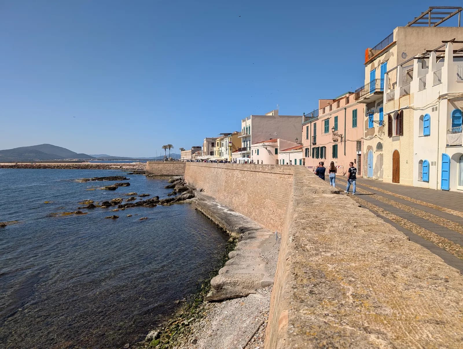 The sea wall of Alghero with waves breaking on rocks below and a row of pastel houses with blue shutters along the walkway
