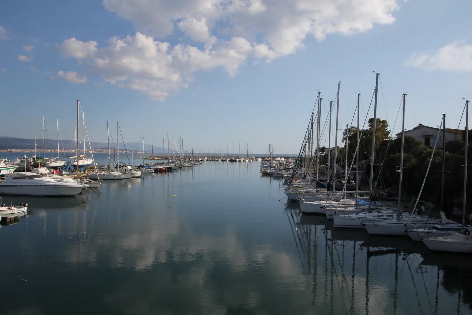 The marina at Fertilia with sailing boats moored on both sides of a still channel