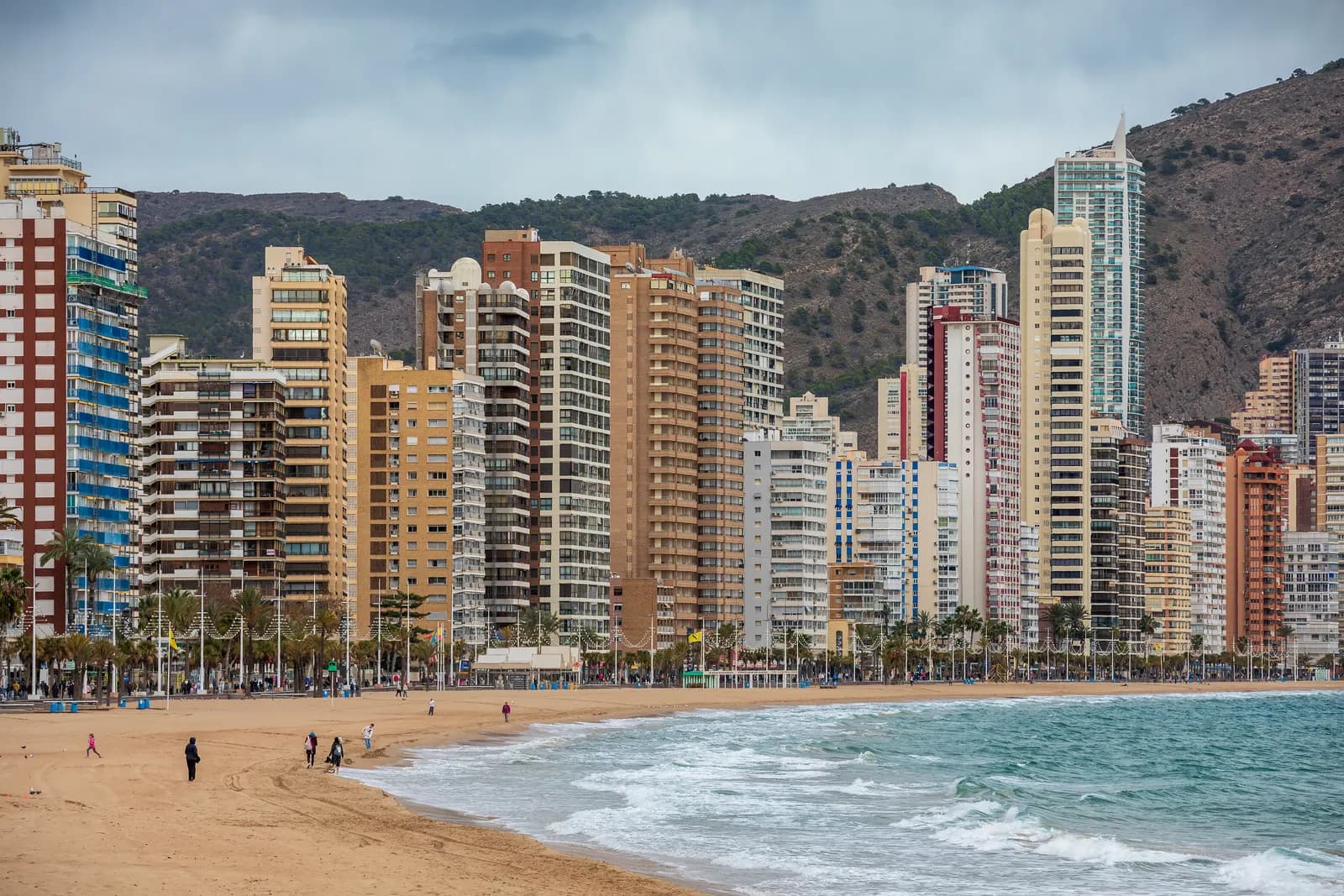 The high-rise apartment towers of Benidorm behind Levante beach, with waves breaking on the sand and hills behind.