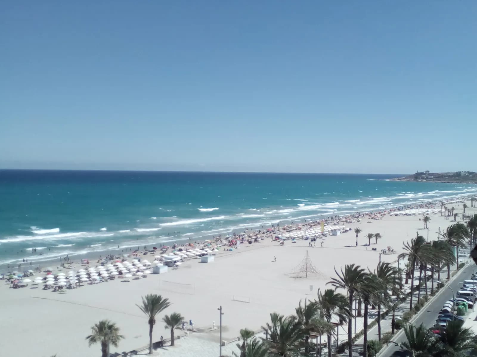 The wide sand of Playa de San Juan seen from above, with parasols, a palm-lined promenade and cars parked along the road.