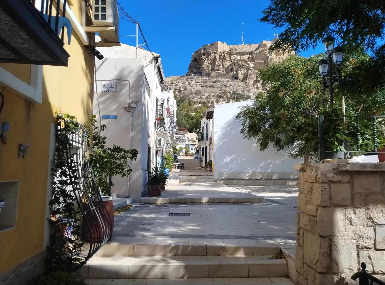 A stepped lane in the Santa Cruz quarter of Alicante with white and yellow houses and plants, and Santa Bárbara castle on the rock above.