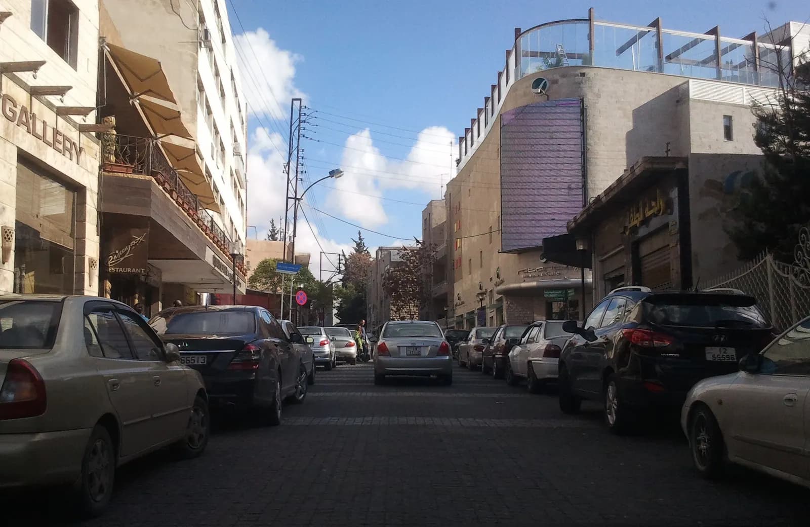Cars parked along both sides of cobbled Rainbow Street in Jabal Amman, between a gallery, a restaurant and stone buildings