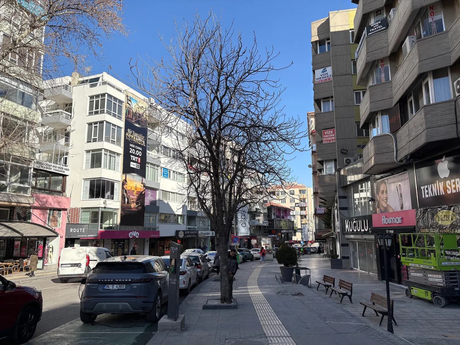 Tunalı Hilmi Caddesi in Ankara with cars parked along the kerb, shops at street level, benches and a bare tree on the pavement