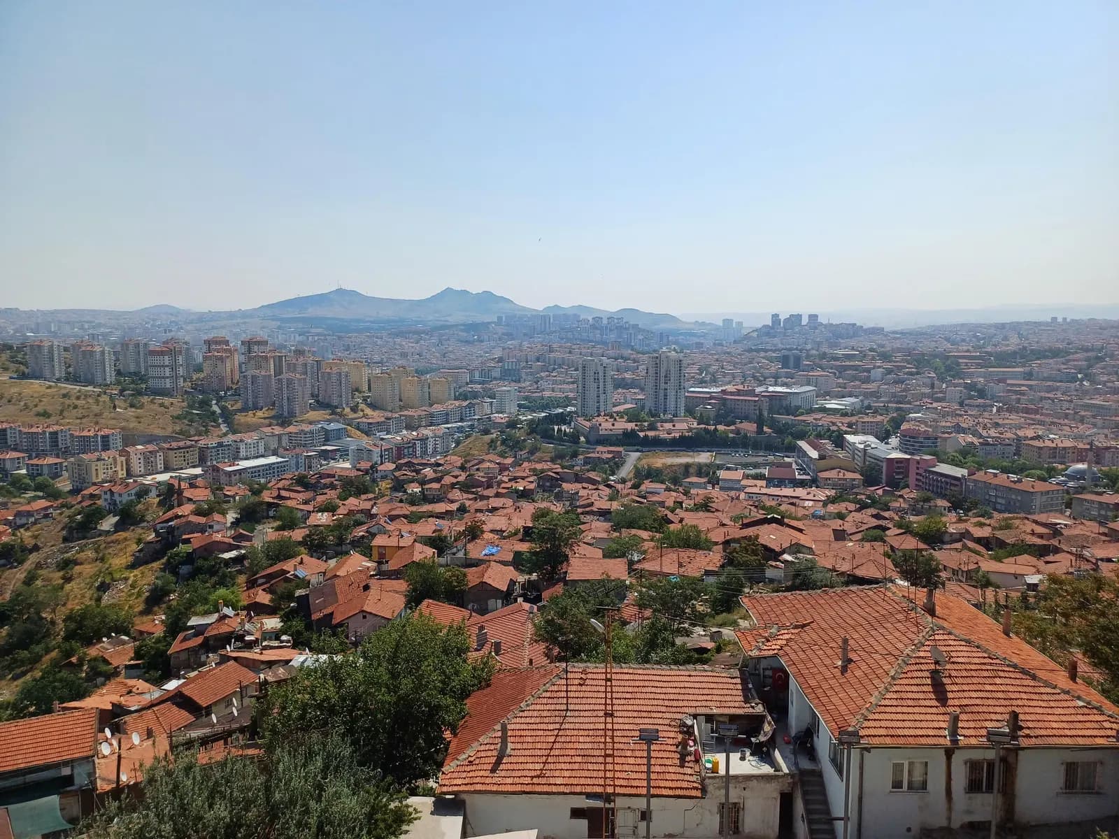 Red tiled roofs of old houses below Ankara Castle, with apartment towers and hills across the city behind