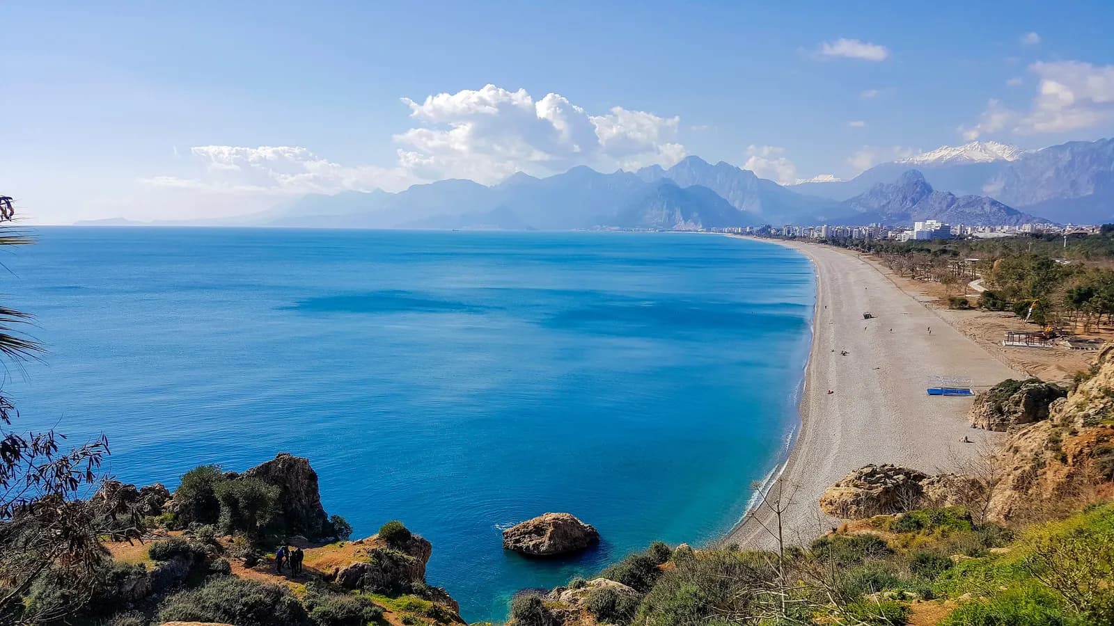 Konyaaltı beach curving west along a turquoise bay below the cliffs of Antalya, with apartment blocks and a range of mountains behind