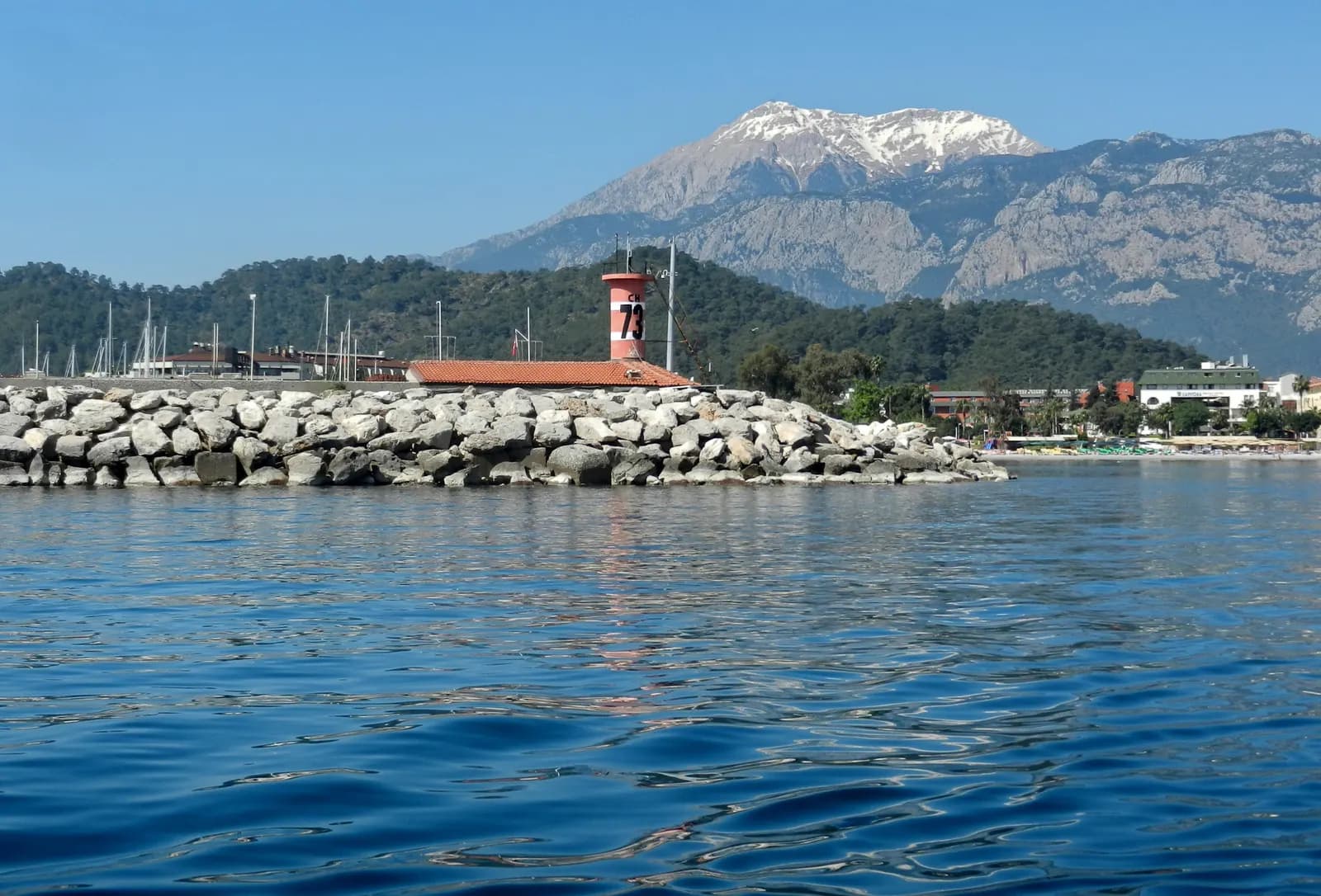 The rock breakwater and harbour light at the entrance to Kemer marina, with pine hills and a snow-capped mountain behind