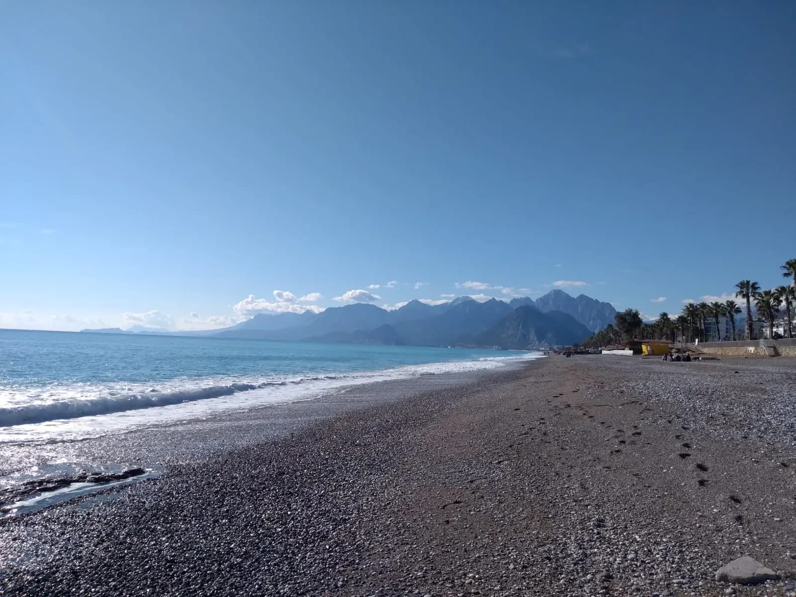 The pebble shore of Konyaaltı beach with small waves, palm trees along the promenade and mountains across the bay