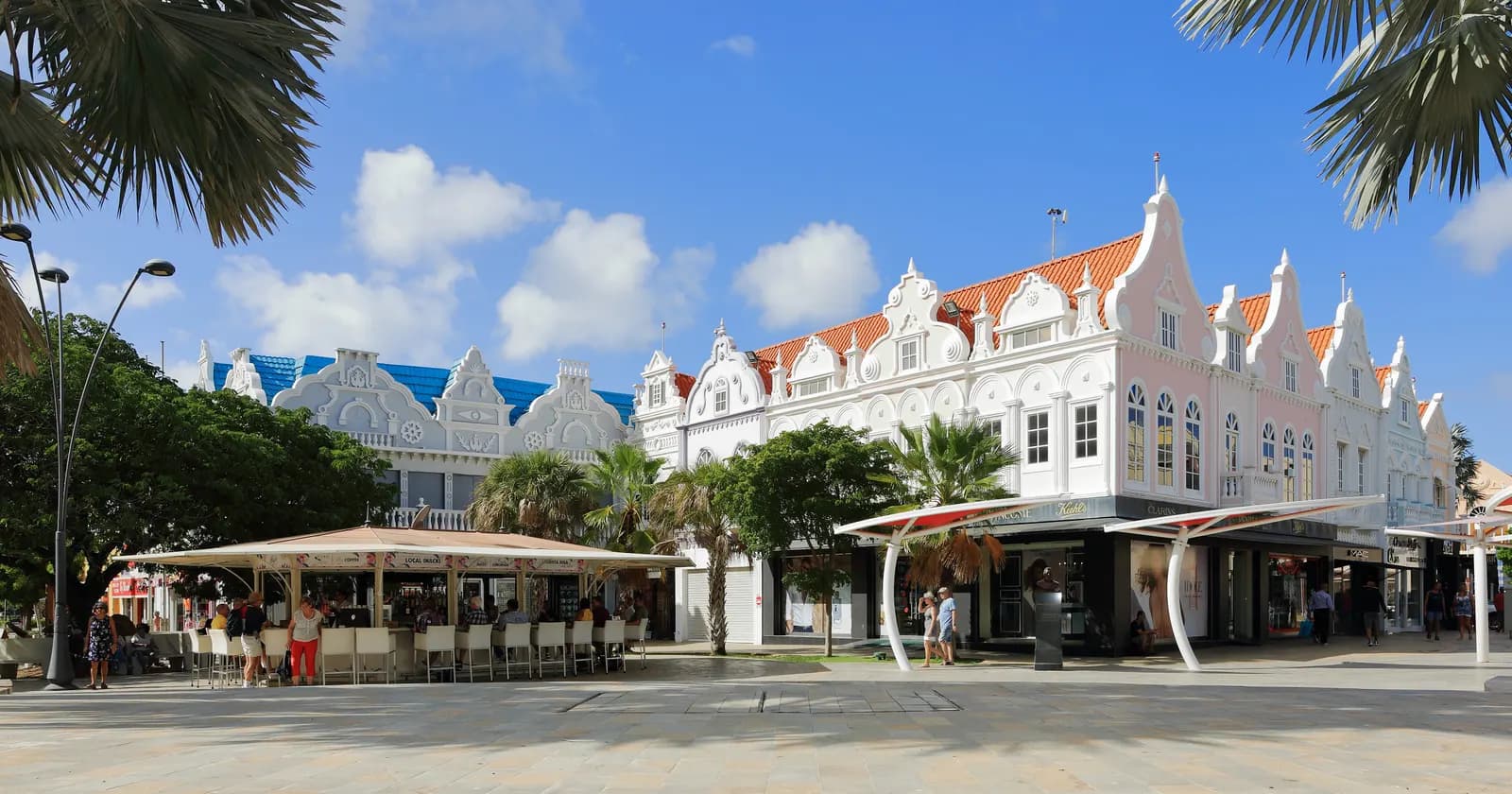 White colonial-style buildings with ornate gables and orange roofs around Plaza Daniel Leo in Oranjestad, with palm trees and café tables